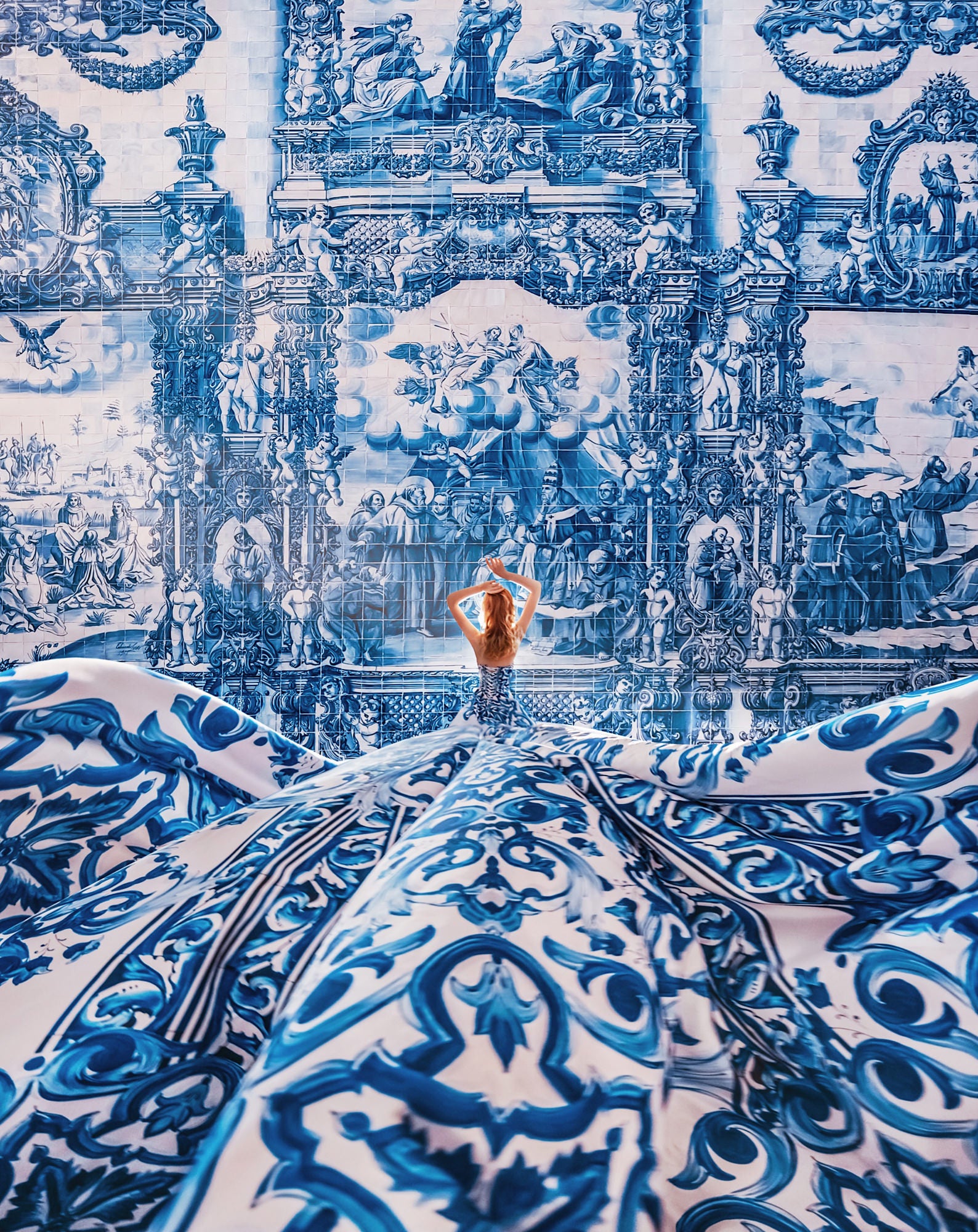 A woman stands before an elaborate blue-and-white tiled mural, framed by sweeping ceramic waves and ornate painted scenes, figures, and floral motifs. Photo by Kristina Makeeva. Alpha 7R IV. 24-70mm f/2.8 G Master.