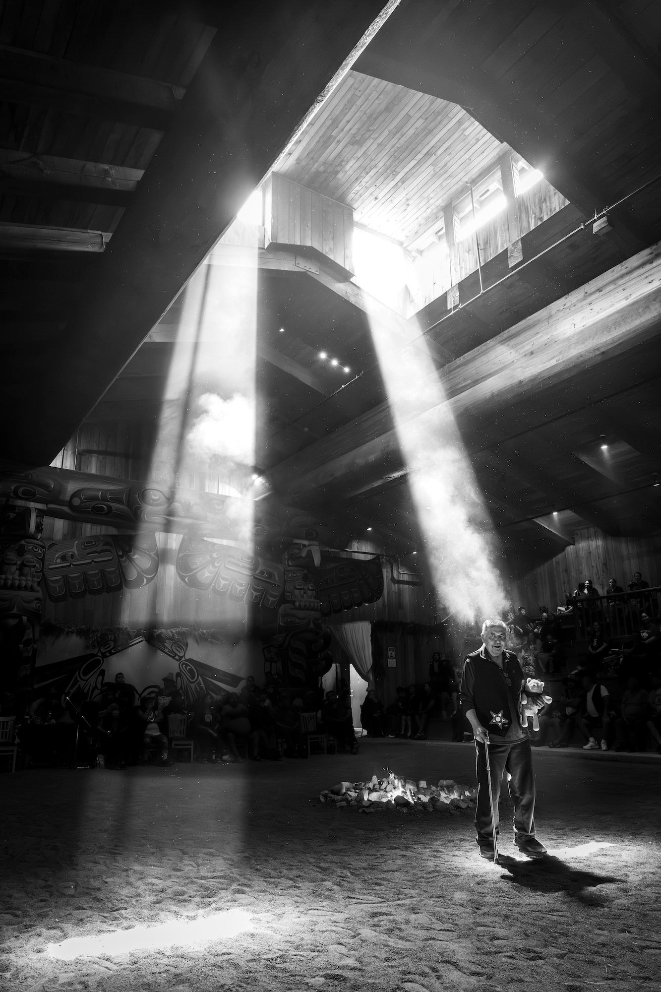 Shafts of light pierce the smoky interior of a wooden longhouse as a man stands near a small fire, black and white. Photo by Jack Plant. Alpha 1. 24-105mm f/4 G. 1/100-sec., f/4, ISO 4000. Shafts of light pierce the smoky interior of a wooden longhouse as a man stands near a small fire, black and white. Photo by Jack Plant. Alpha 1. 24-105mm f/4 G. 1/100-sec., f/4, ISO 4000.