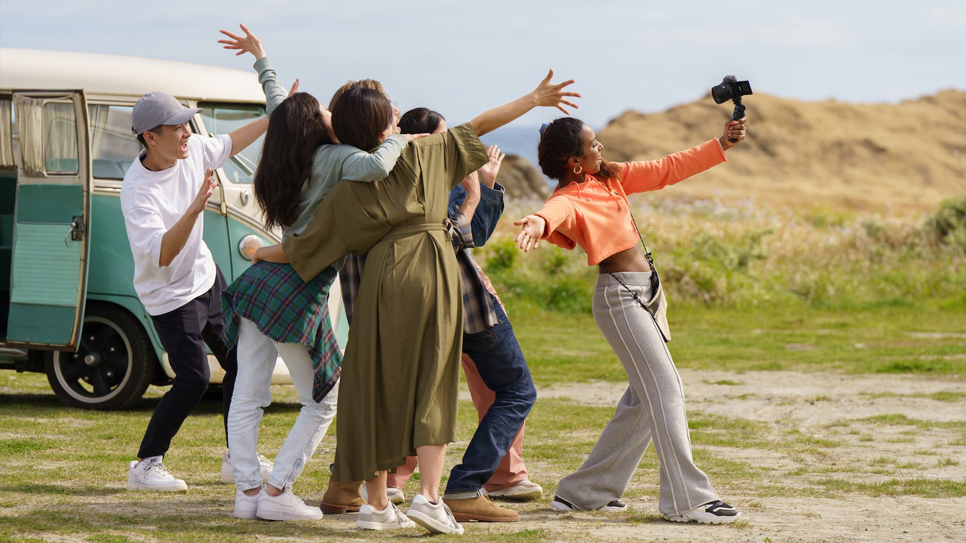 A woman holds a vlogging camera up to herself and a group of friends behind her.