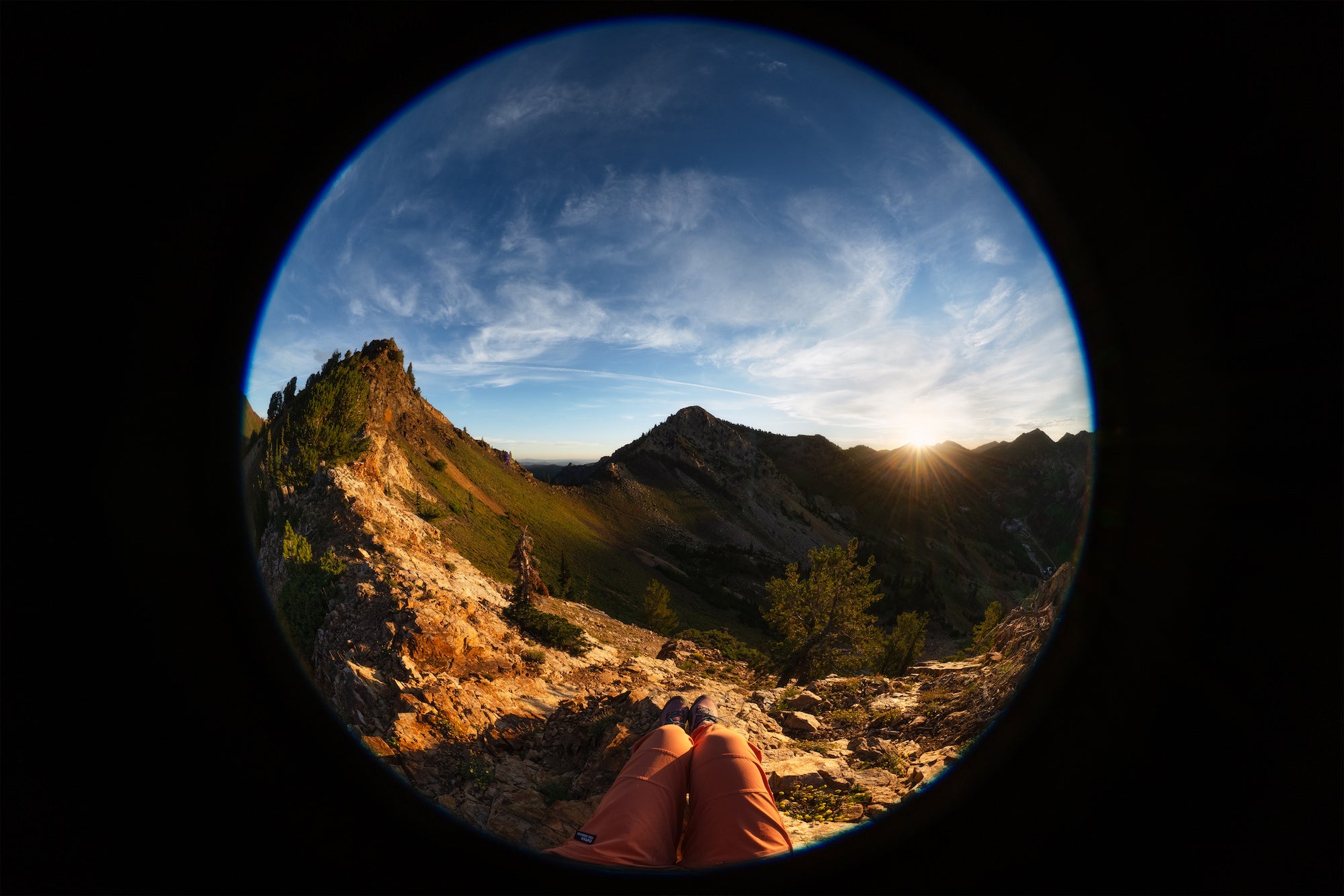 Circular fisheye view of a person&rsquo;s legs resting on a rocky mountain ridge at sunset, photographed by Autumn Schrock.