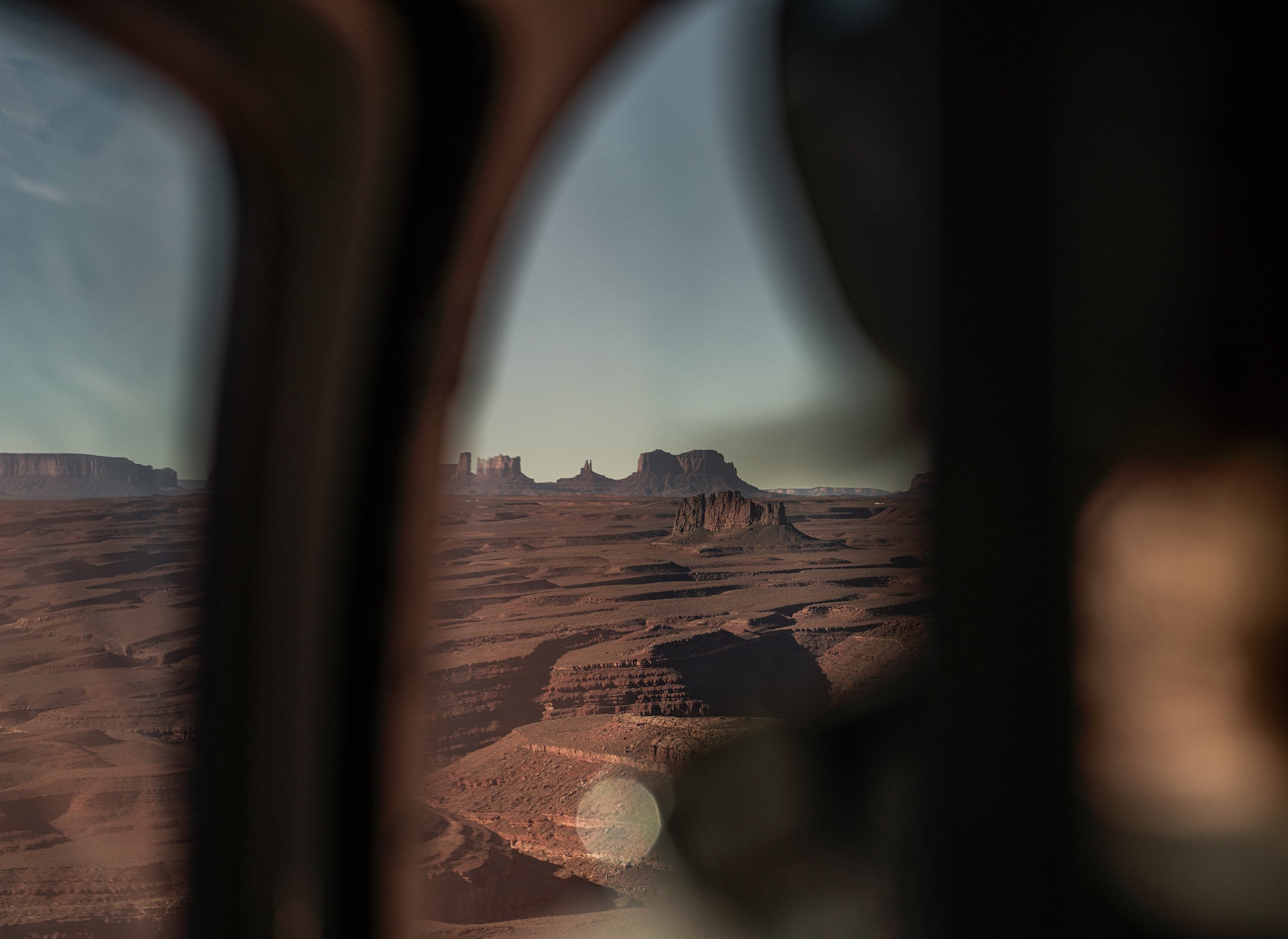 View of distant red-rock buttes and mesas through a helicopter window, framed by the aircraft&rsquo;s dark interior. Photograph by Sydney Smolla.
