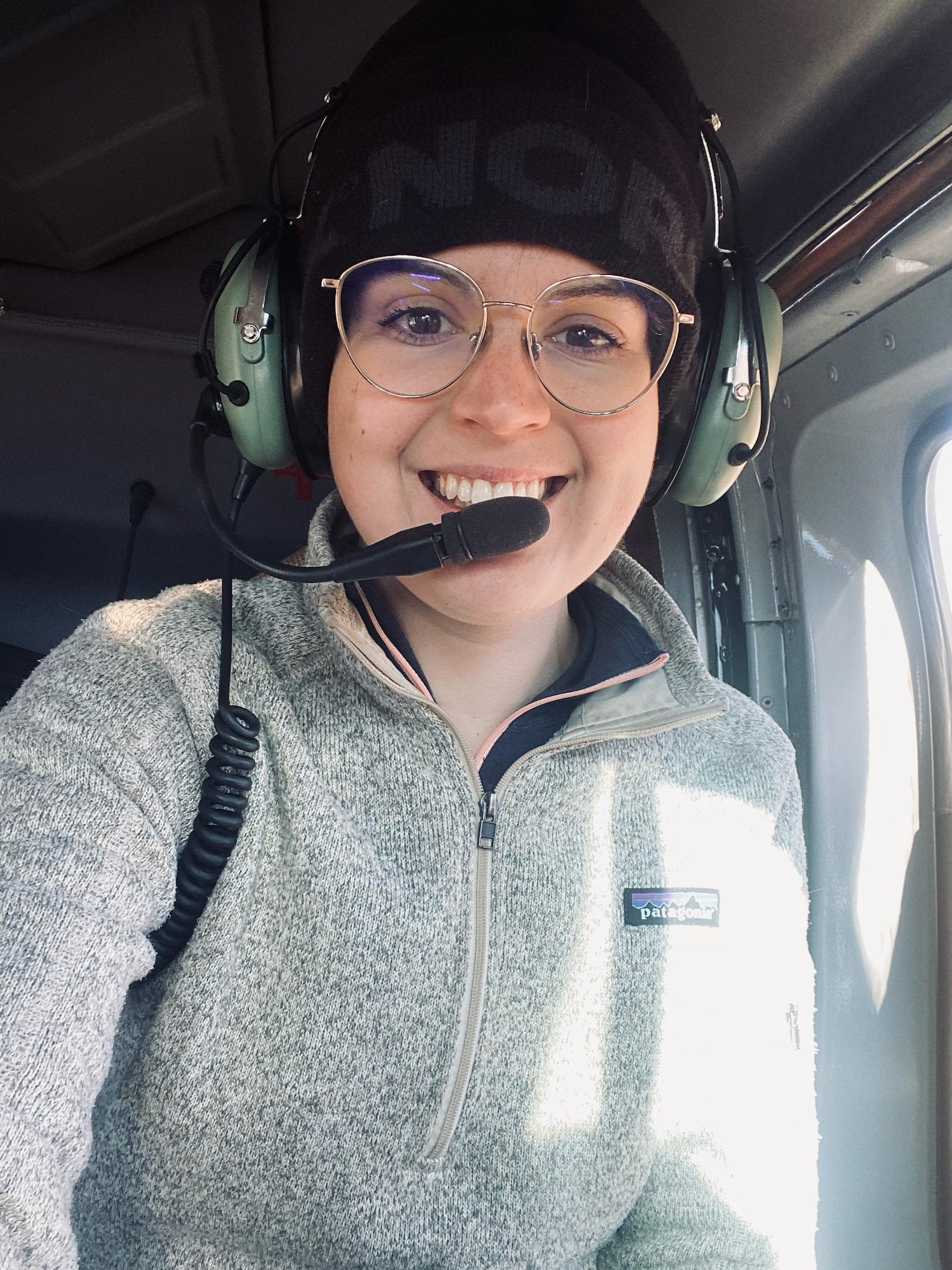 Sydney Smolla smiles inside a helicopter, wearing glasses, a black beanie, and a green aviation headset. Photograph by Sydney Smolla.