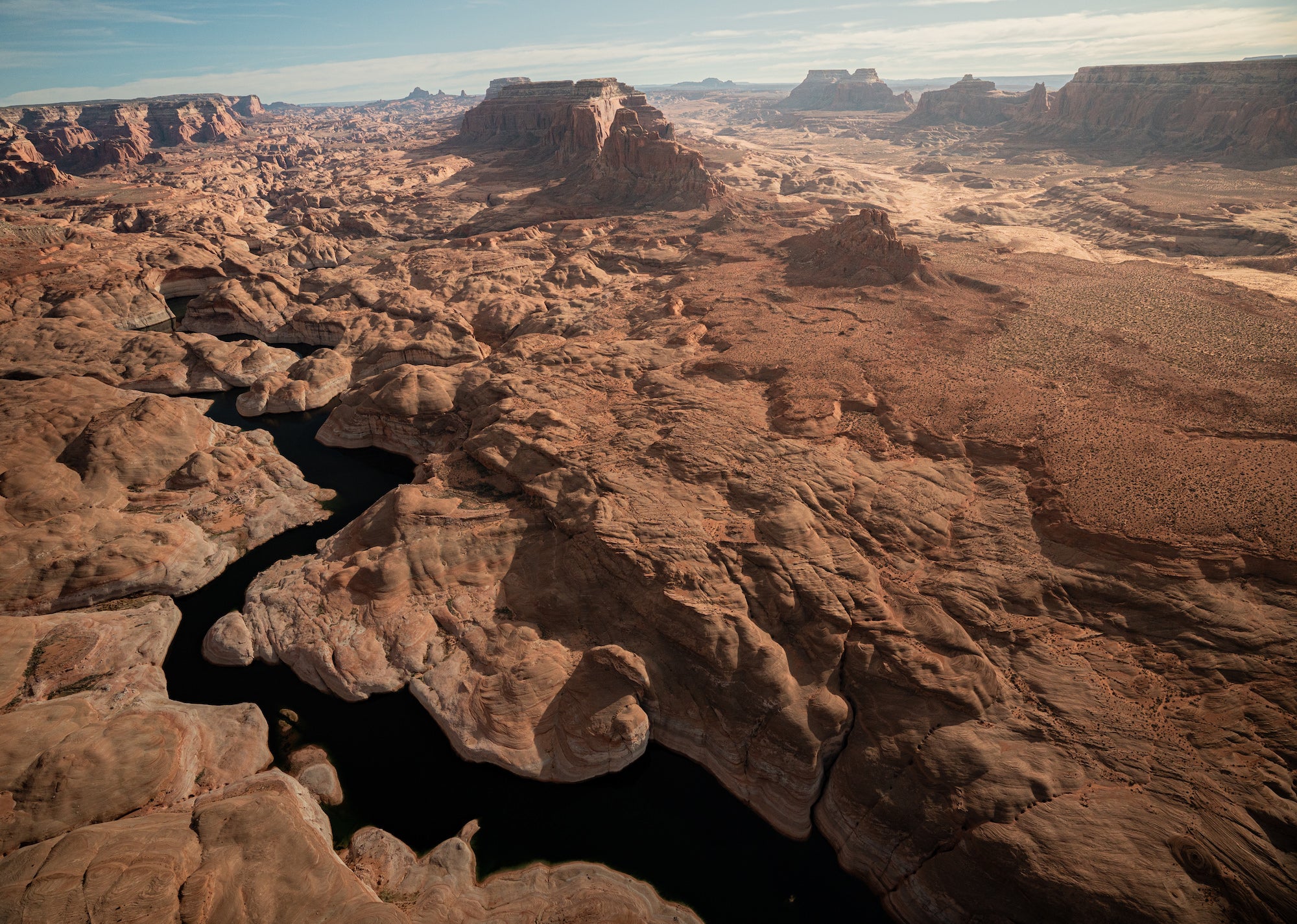 Aerial view of a narrow dark-water channel winding through sculpted sandstone formations and a vast desert landscape. Photograph by Sydney Smolla.