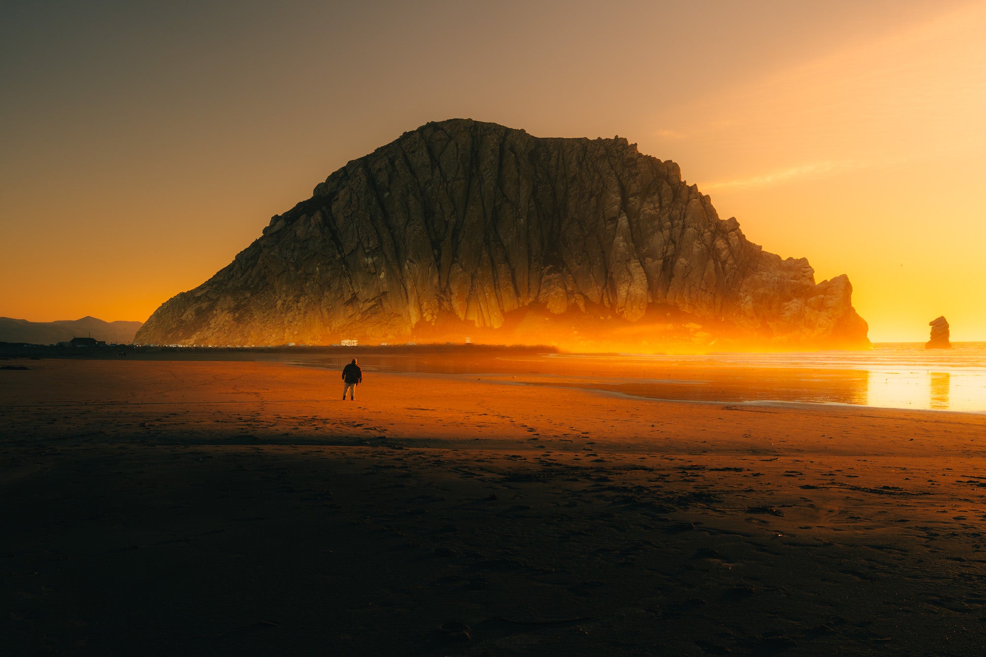 Massive seaside rock at sunset with a lone figure on the glowing beach.