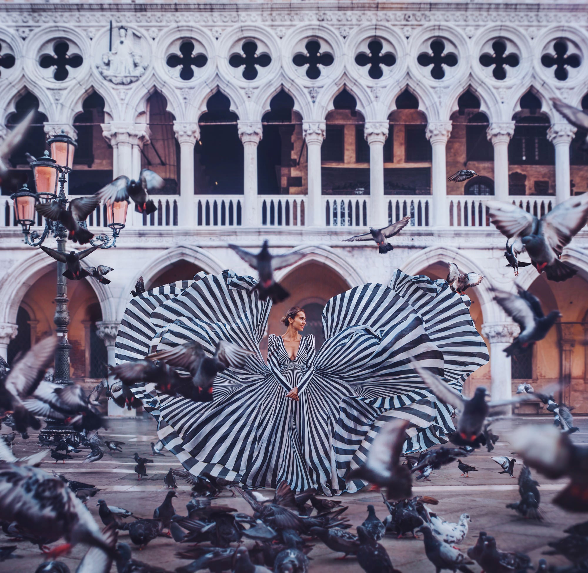 A woman in a dramatic black-and-white striped gown stands in a grand arched courtyard as pigeons gather and fly around her. Photo by Kristina Makeeva. Alpha 7R IV. 35mm f/1.4 G Master.