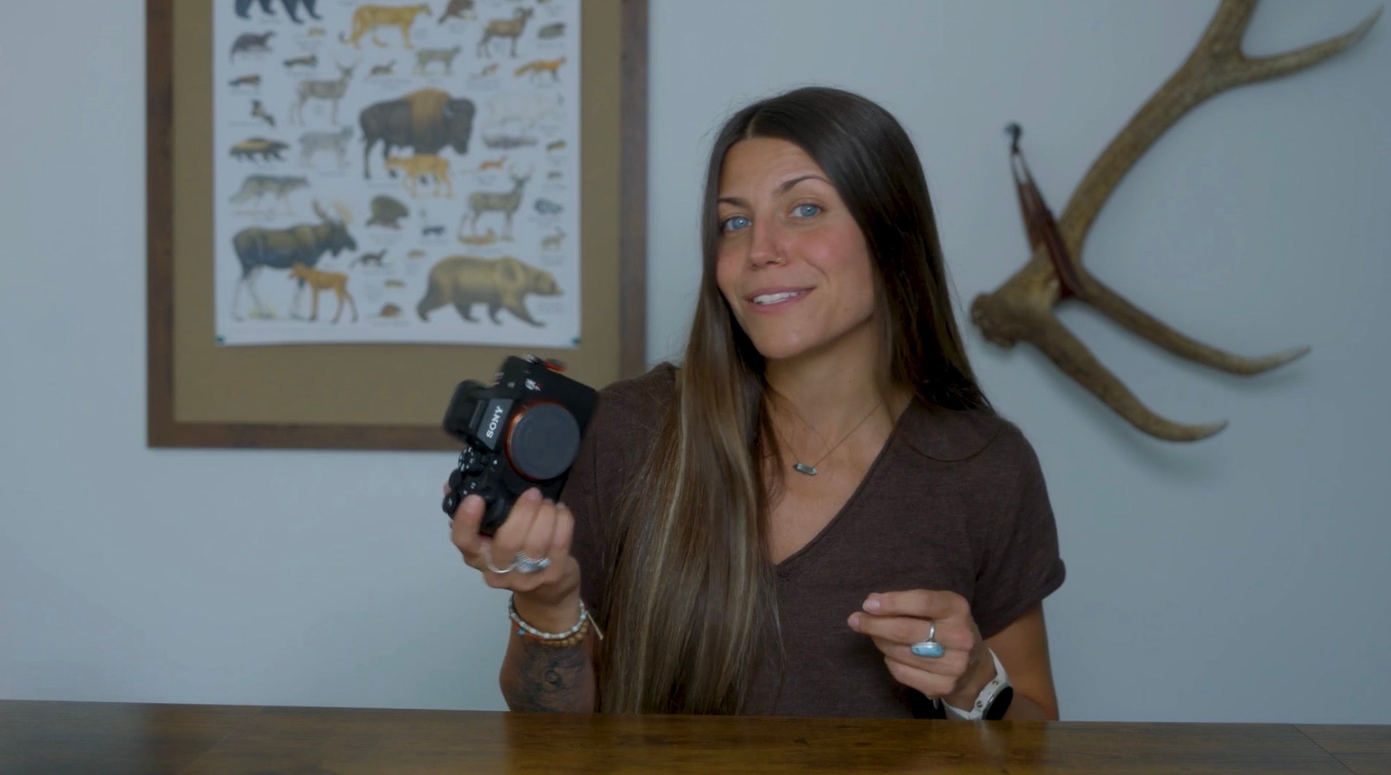 Hannah Rheaume holds up a Sony Alpha mirrorless camera body at a wooden table, antlers and wildlife poster behind her.