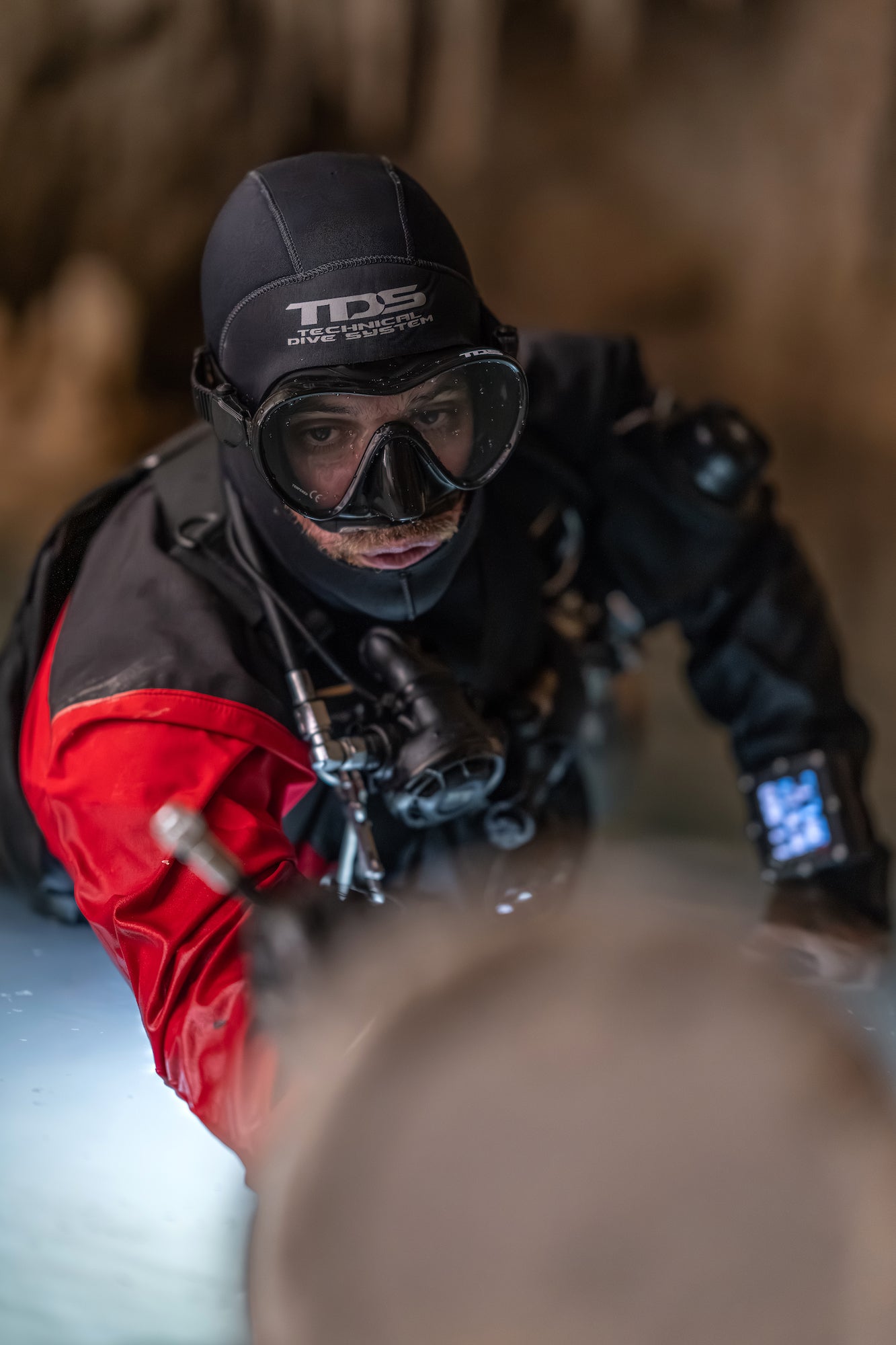Underwater portrait of photographer &Aacute;lvaro &ldquo;Mekan&rdquo; Herrero wearing a black wetsuit, dive mask, and red drysuit while holding a camera inside a dimly lit cavern. Soft light catches his face and equipment against the shadowy rock walls.