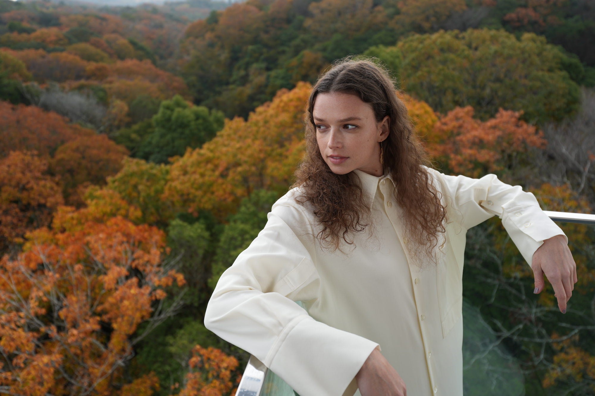 A woman in a white blouse poses before a hillside of green, orange and red autumn foliage. A woman in a white blouse poses before a hillside of green, orange and red autumn foliage.