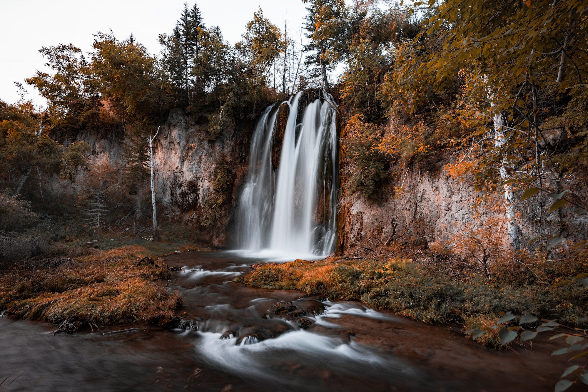 Long-exposure photograph of a waterfall flowing into a rocky stream surrounded by autumn foliage. Long-exposure photograph of a waterfall flowing into a rocky stream surrounded by autumn foliage.