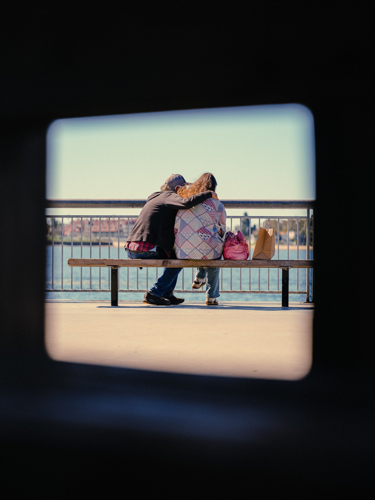Candid couple sharing a bench on a waterfront promenade, framed through a window, documentary street photo.