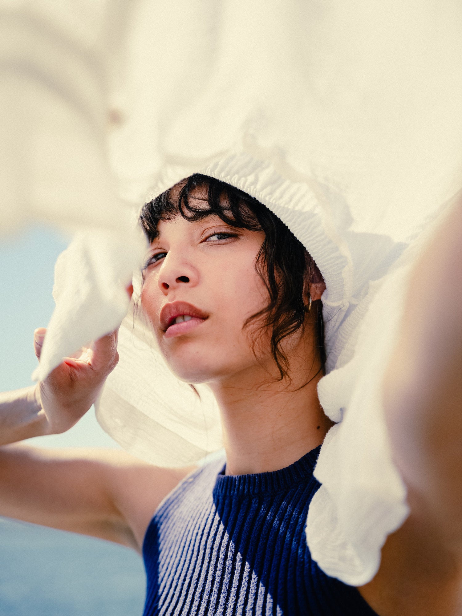 Close‑up portrait framed by a flowing white hat, bright seaside light, editorial beach style.