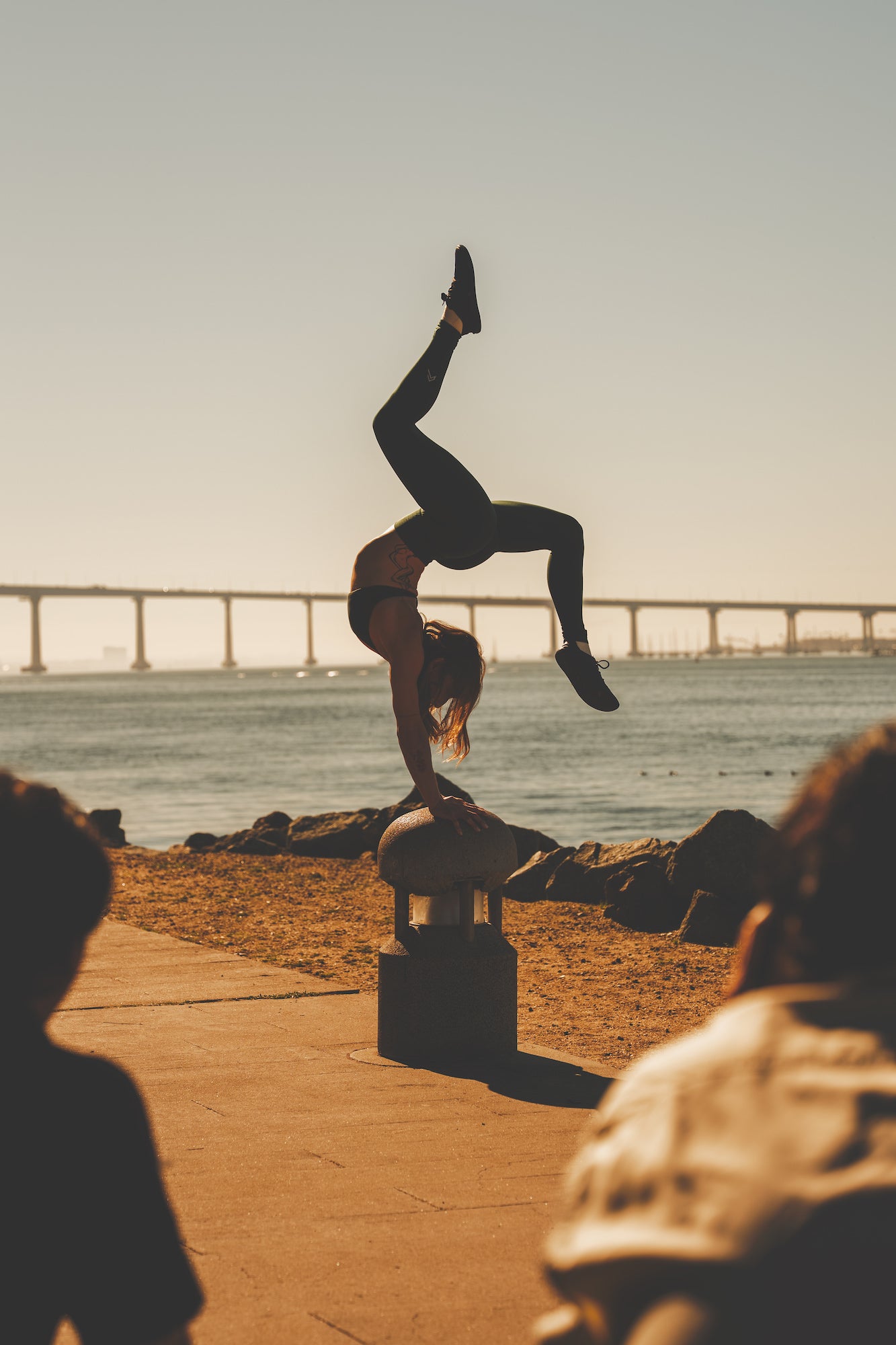 Acrobat performing a one‑hand handstand on a bollard with a long arched bridge over the bay in the background, sunset fitness shot.