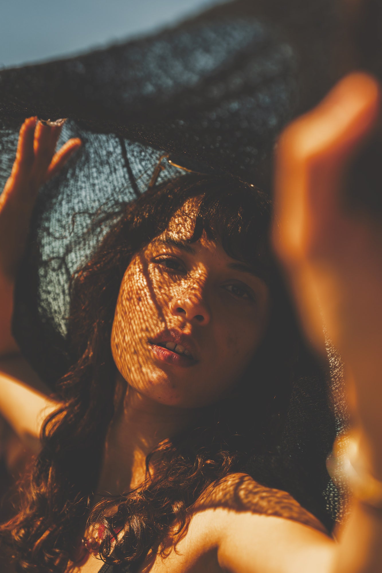 Artistic close‑up portrait with dappled mesh shadows across the face in warm sunset light, coastal editorial style.