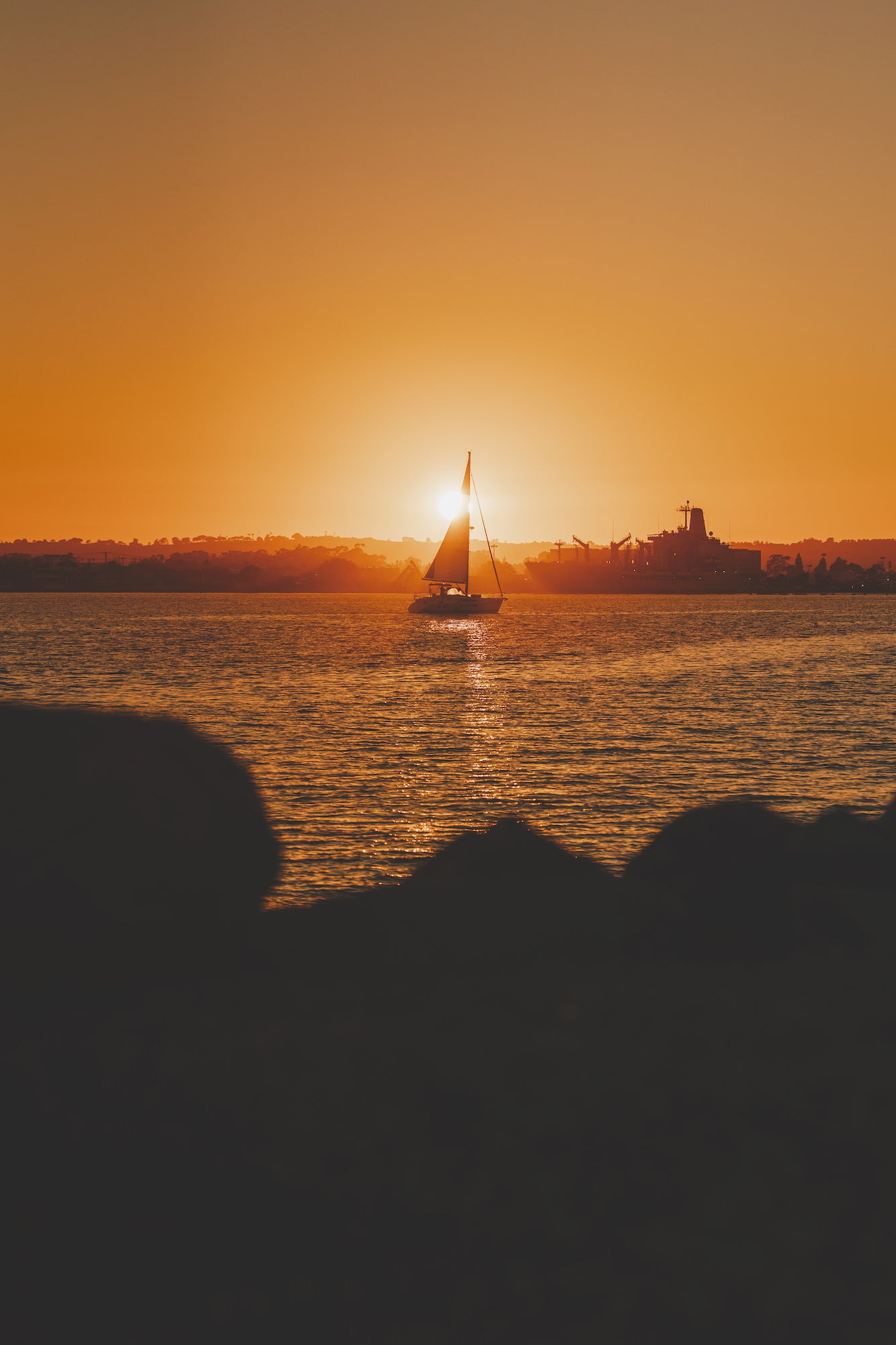 Sailboat silhouette at vivid orange sunset on the bay with distant skyline, San Diego seascape photography.
