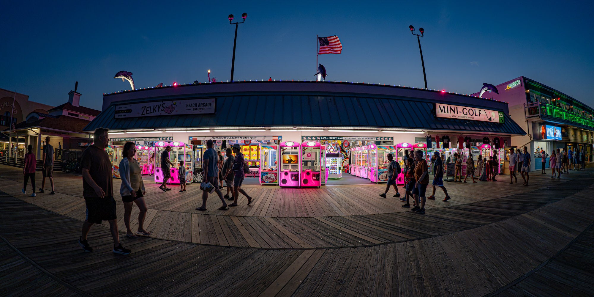 People walk past a brightly lit arcade on a boardwalk at dusk, photographed by Drew Geraci.