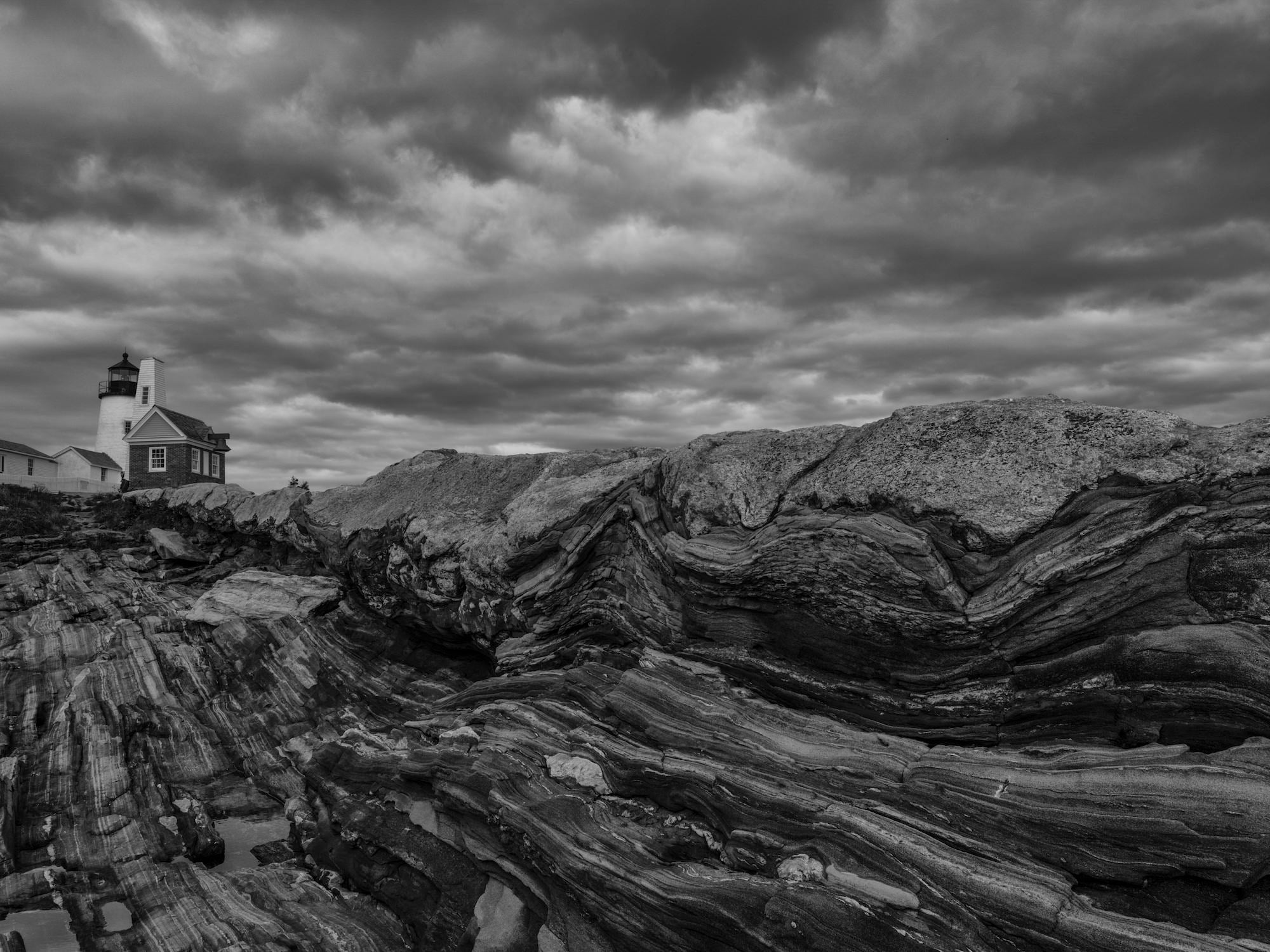 Black-and-white lighthouse scene with dramatic clouds above layered, striated coastal rock formations.