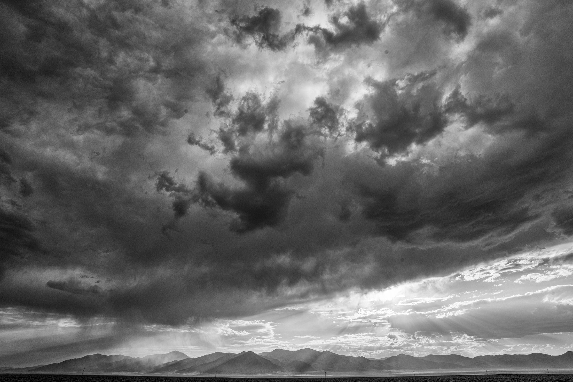 Black-and-white storm clouds with sun rays breaking over a distant mountain range.