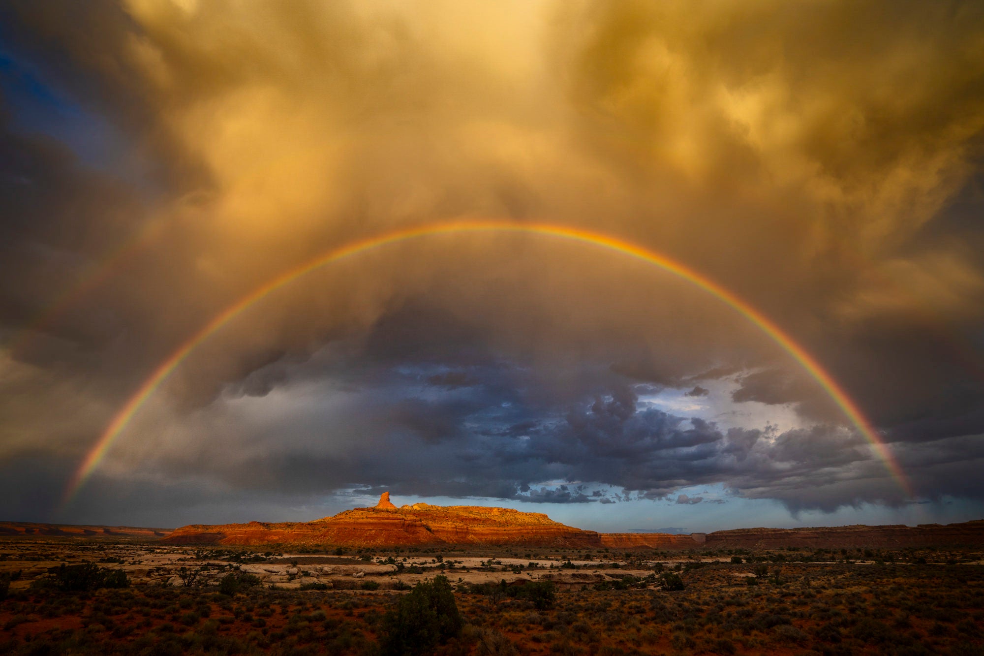 Full rainbow arcing over a sunlit desert mesa under stormy skies at golden hour.