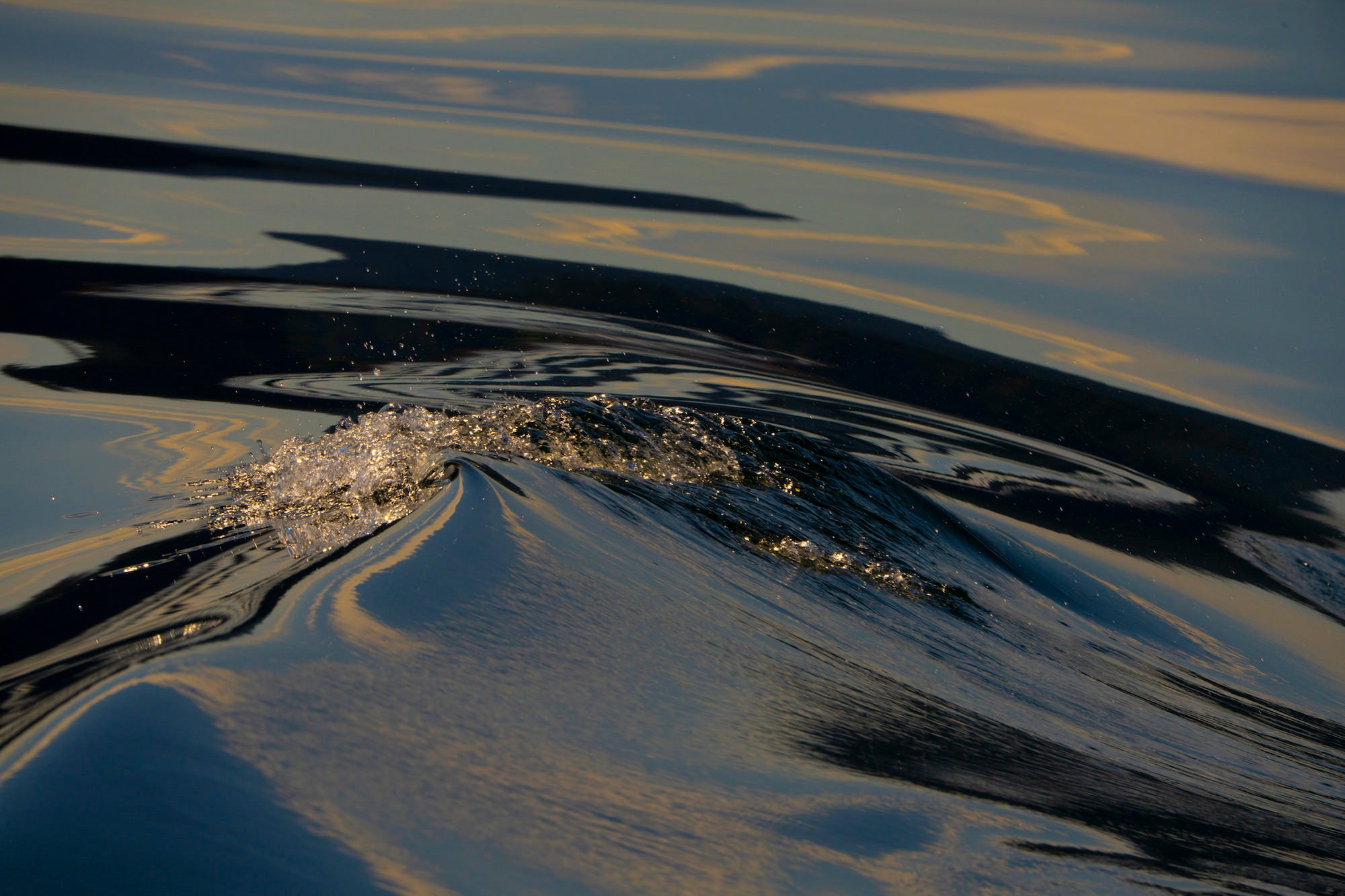 Close-up of a small cresting wave with shimmering highlights and smooth water reflections at sunset.