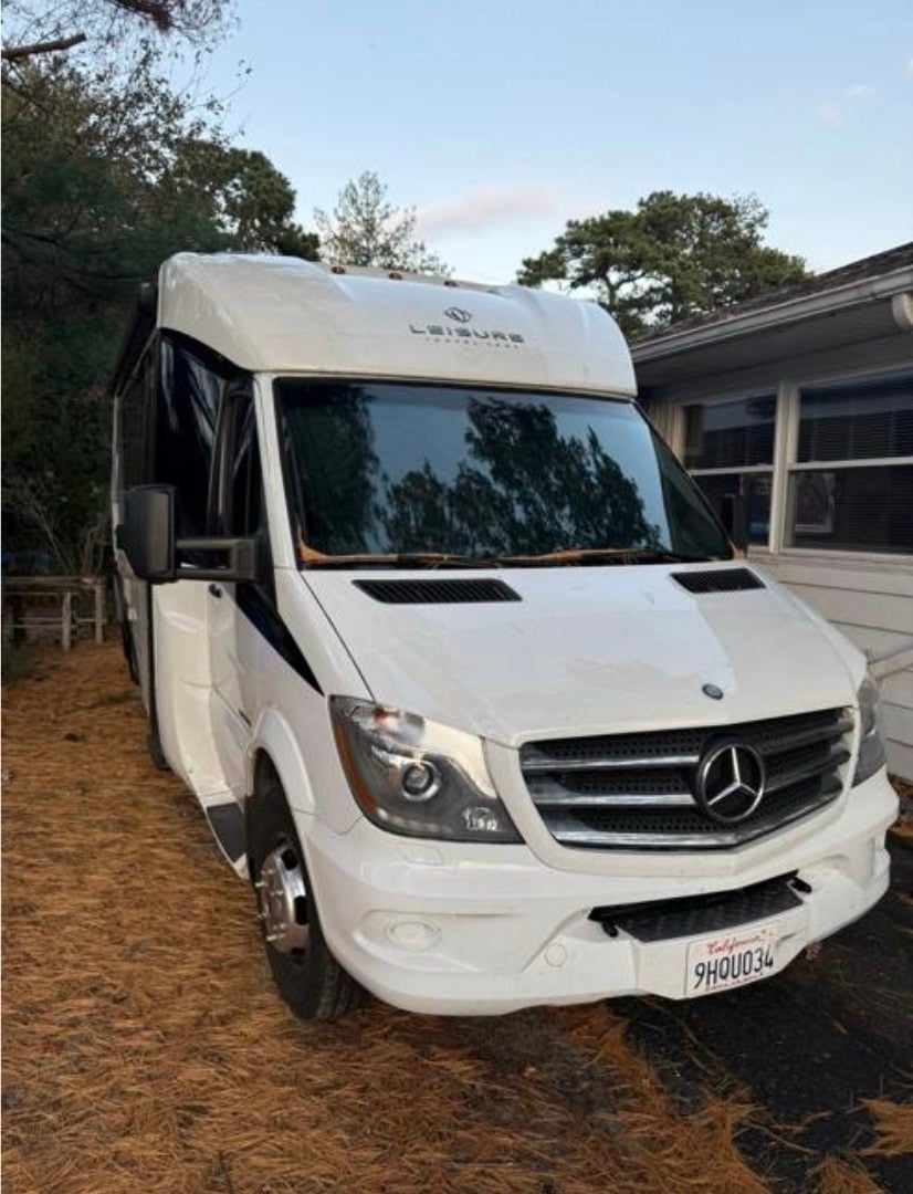 White Mercedes Sprinter-based RV parked beside a house, front view among pine needles.