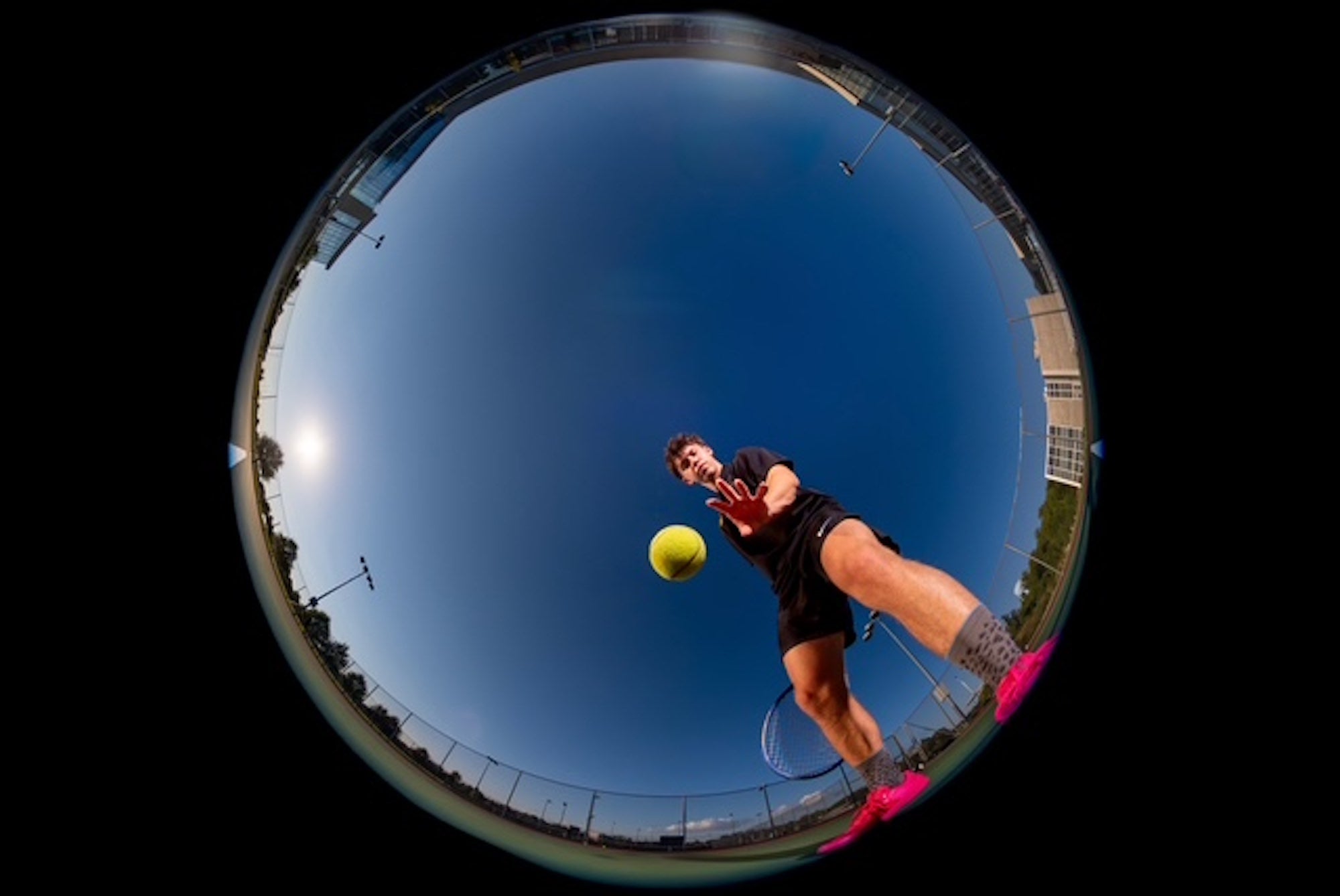A tennis player reaches for a ball in a circular fisheye view from court level, photographed by Brad Deel.