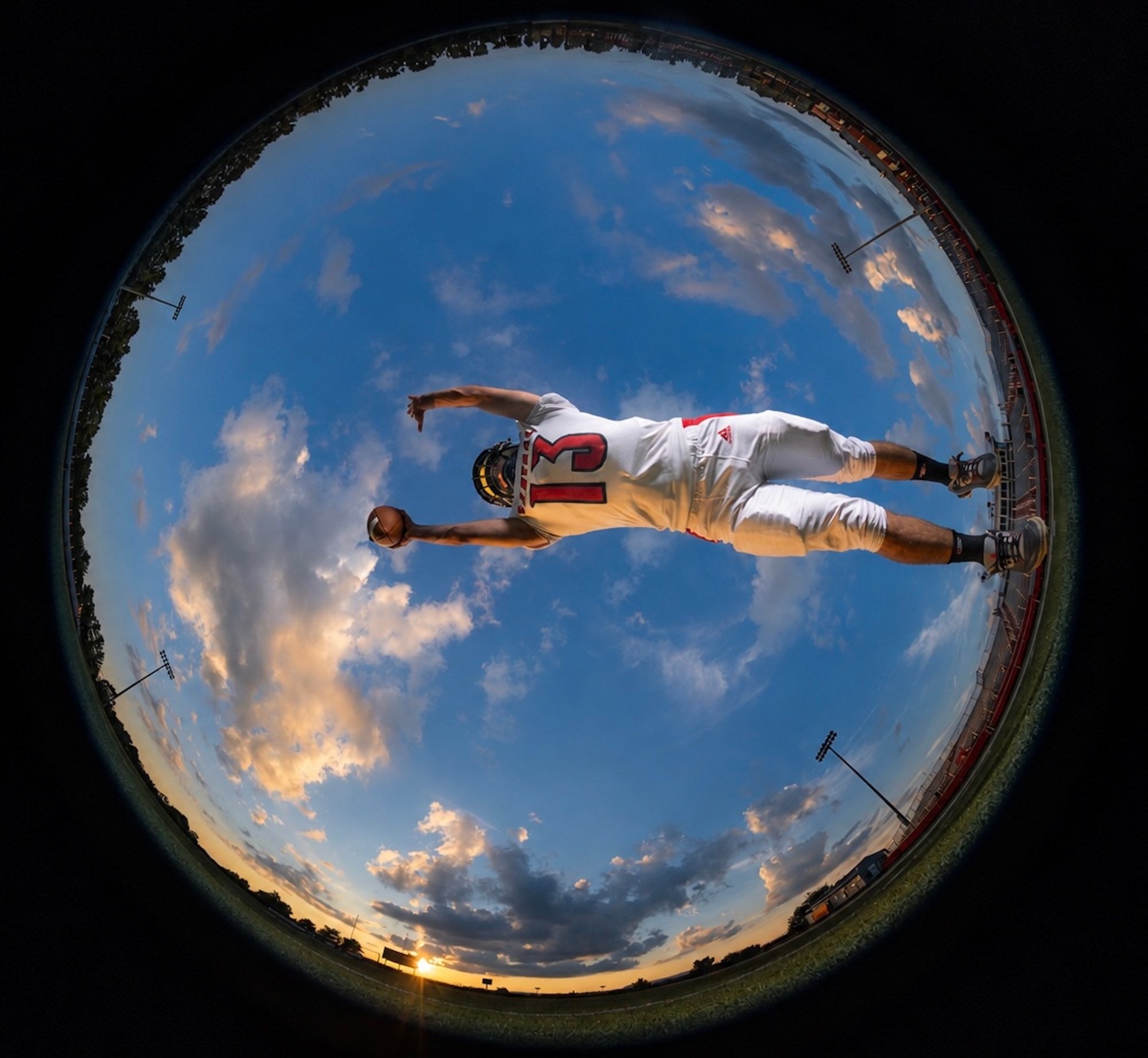 A football player dives with the ball, framed in a circular fisheye view beneath a dramatic sky, photographed by Brad Deel.