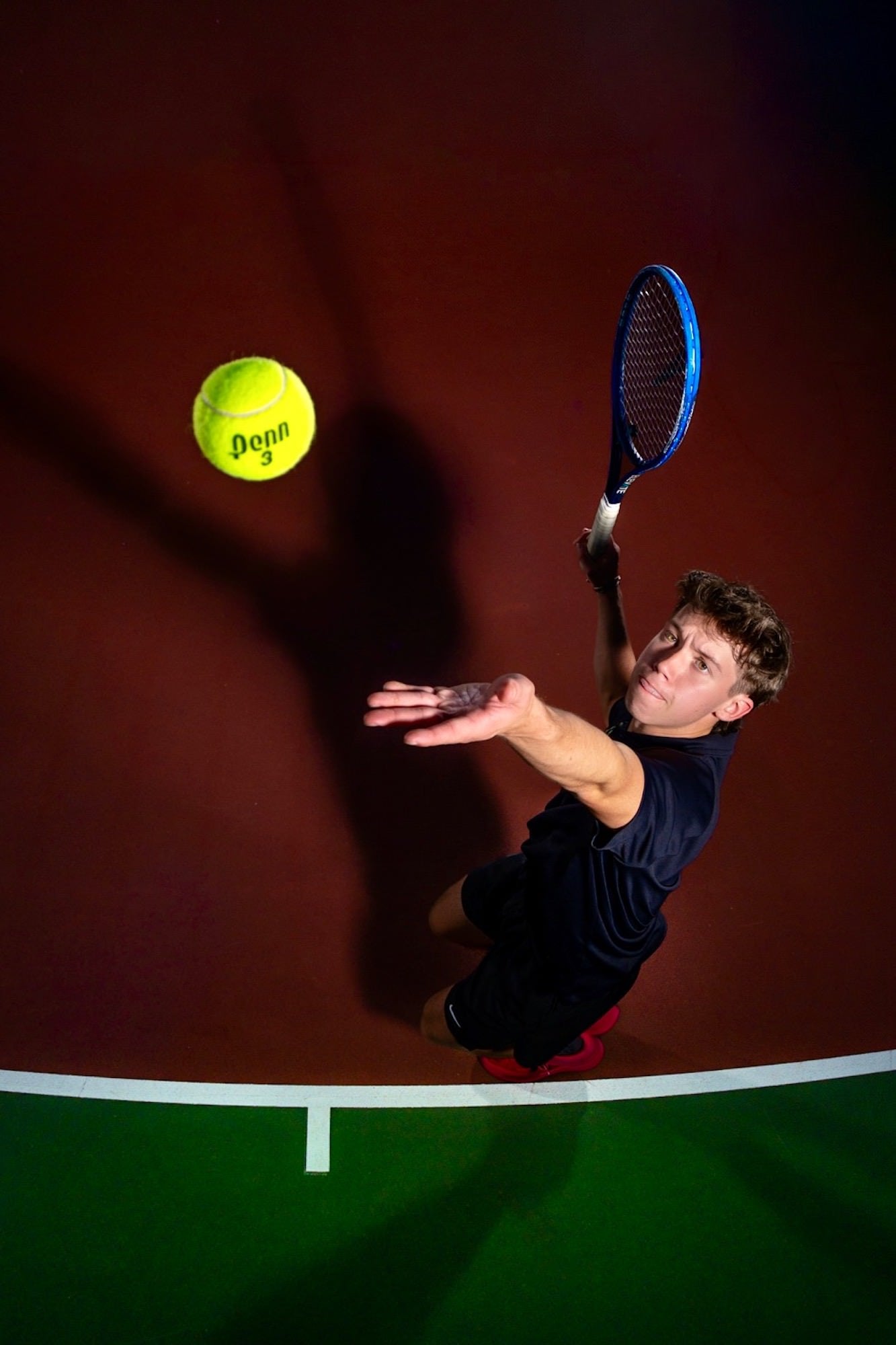 Overhead view of a tennis player serving toward a bright yellow ball on a court, photographed by Brad Deel.
