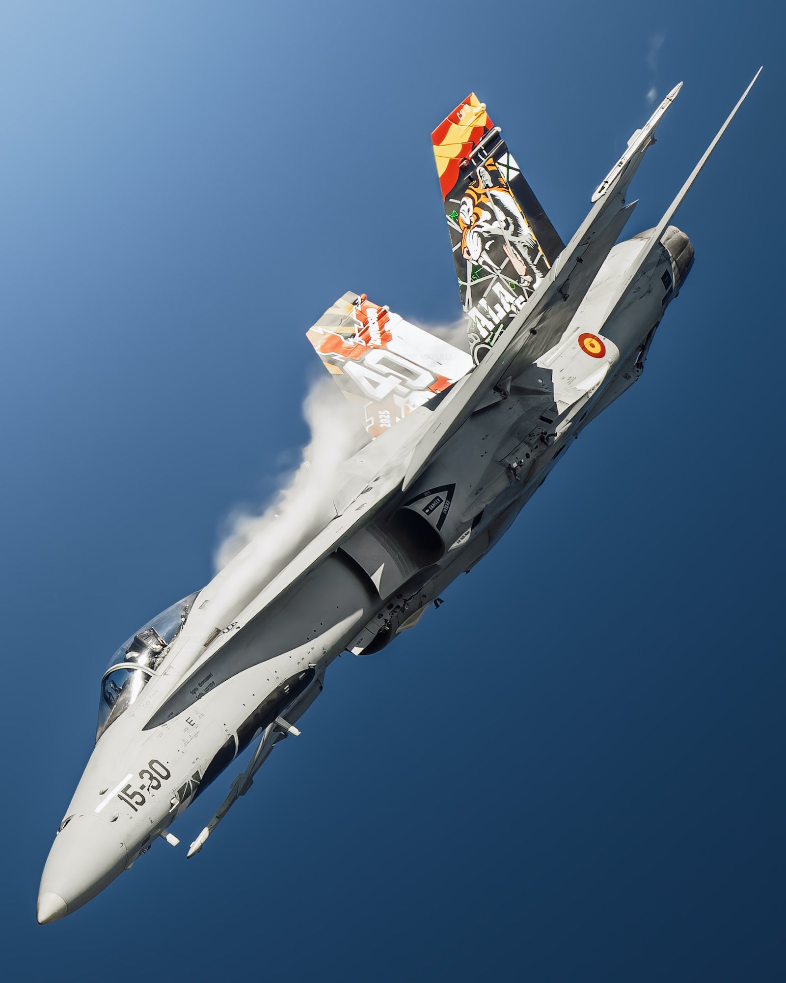 Fighter jet with colorful tail art banks sharply, condensation streaming from the wings against a clear sky.
