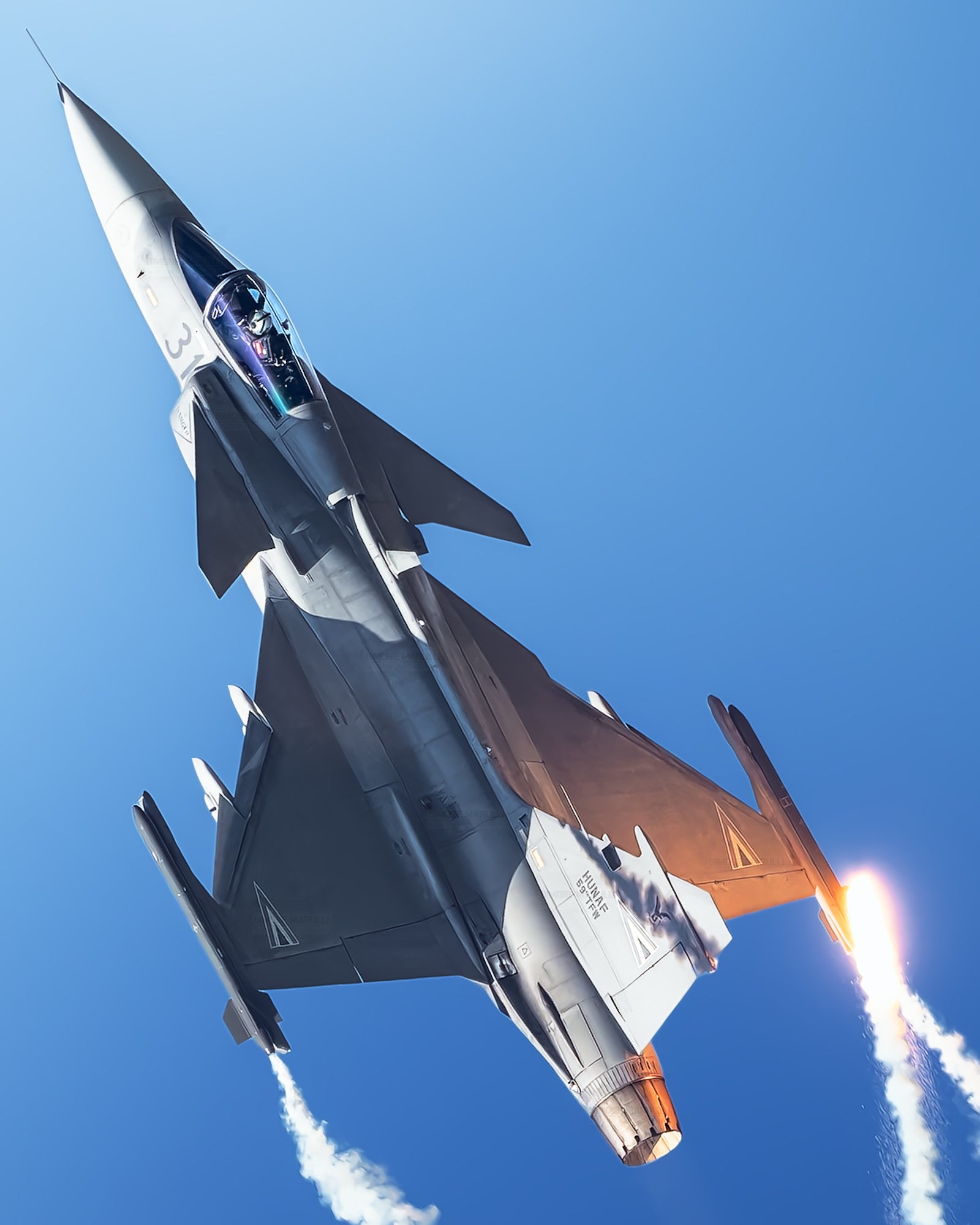 Fighter jet climbs steeply with afterburner lit and white vapor trails against a deep blue sky.