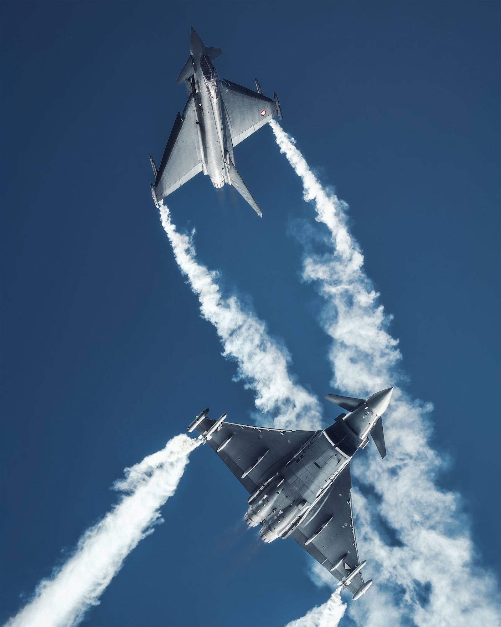Two fighter jets streak upward in formation, leaving long white vapor trails across a clear blue sky.