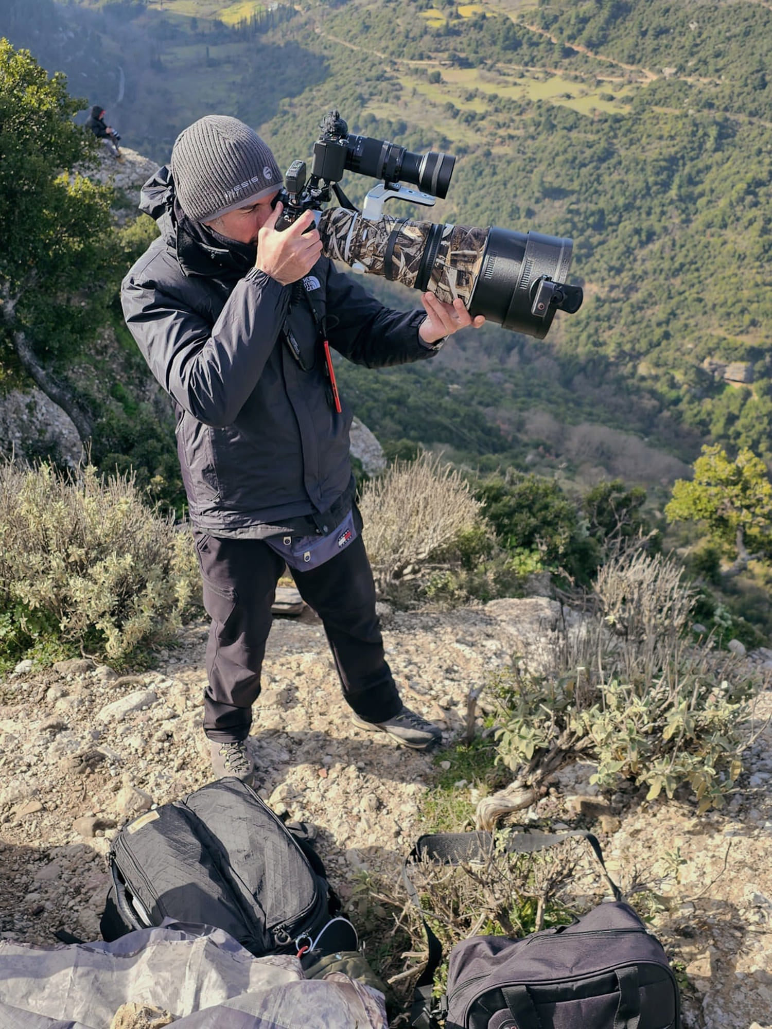 Photographer on a rocky hillside aims a long‑lens camera rig with a second body mounted on top; valley landscape behind.