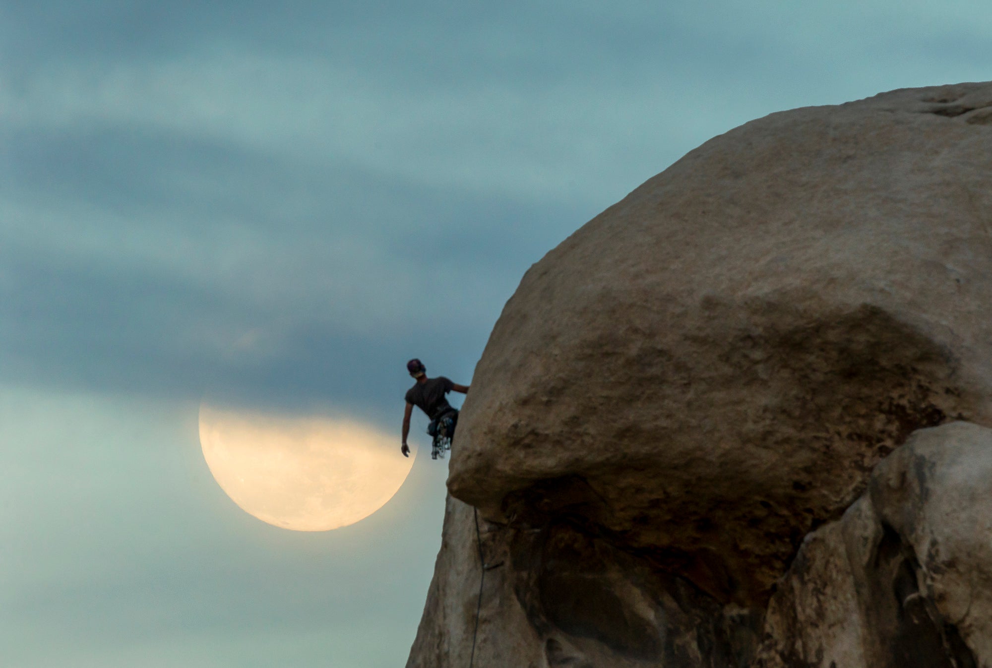 Rock climber on a sandstone dome at dusk with a large pale moon rising behind thin clouds. Rock climber on a sandstone dome at dusk with a large pale moon rising behind thin clouds.