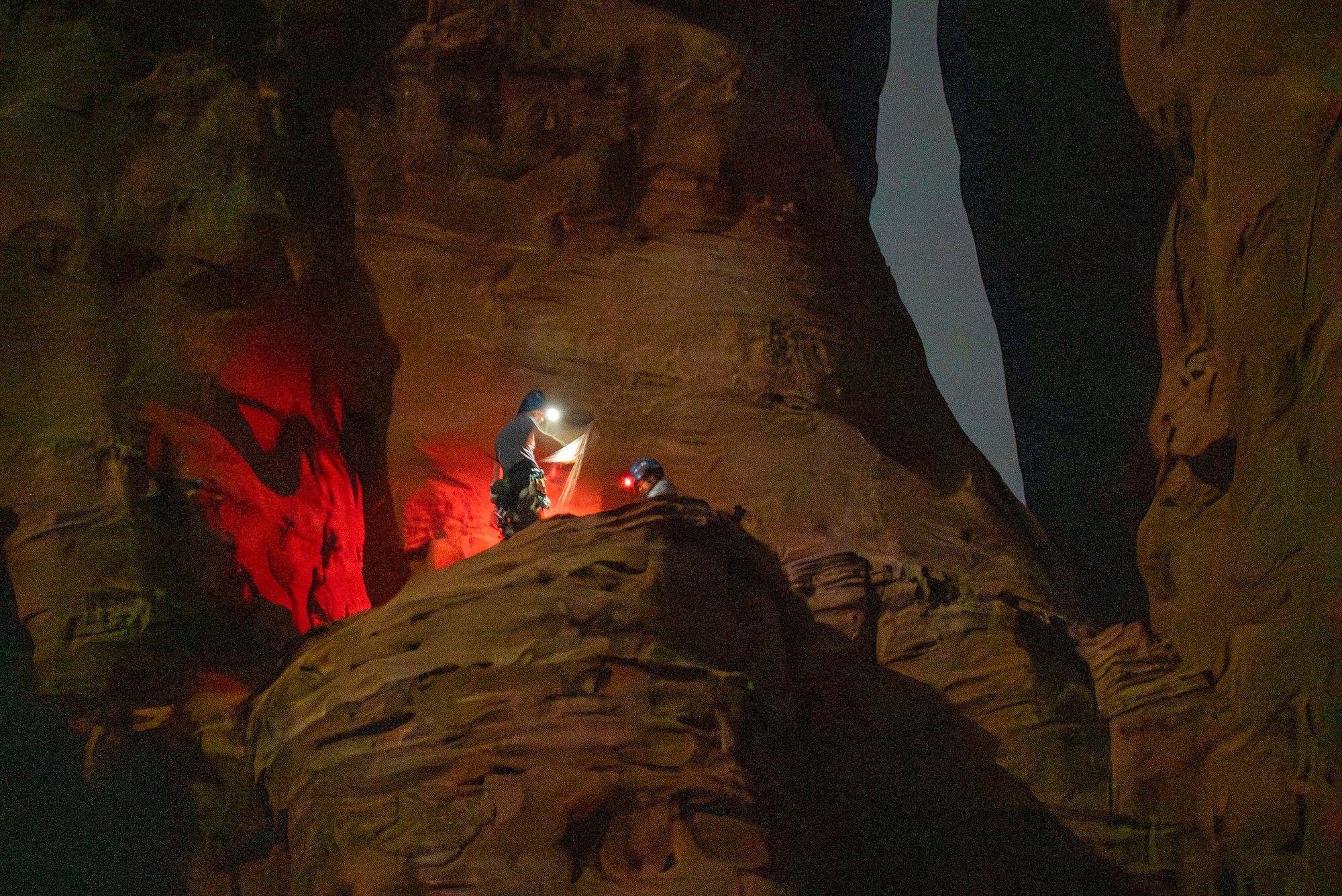 Nighttime ledge scene with climbers using headlamps and a reflective panel amid deep red sandstone walls.
