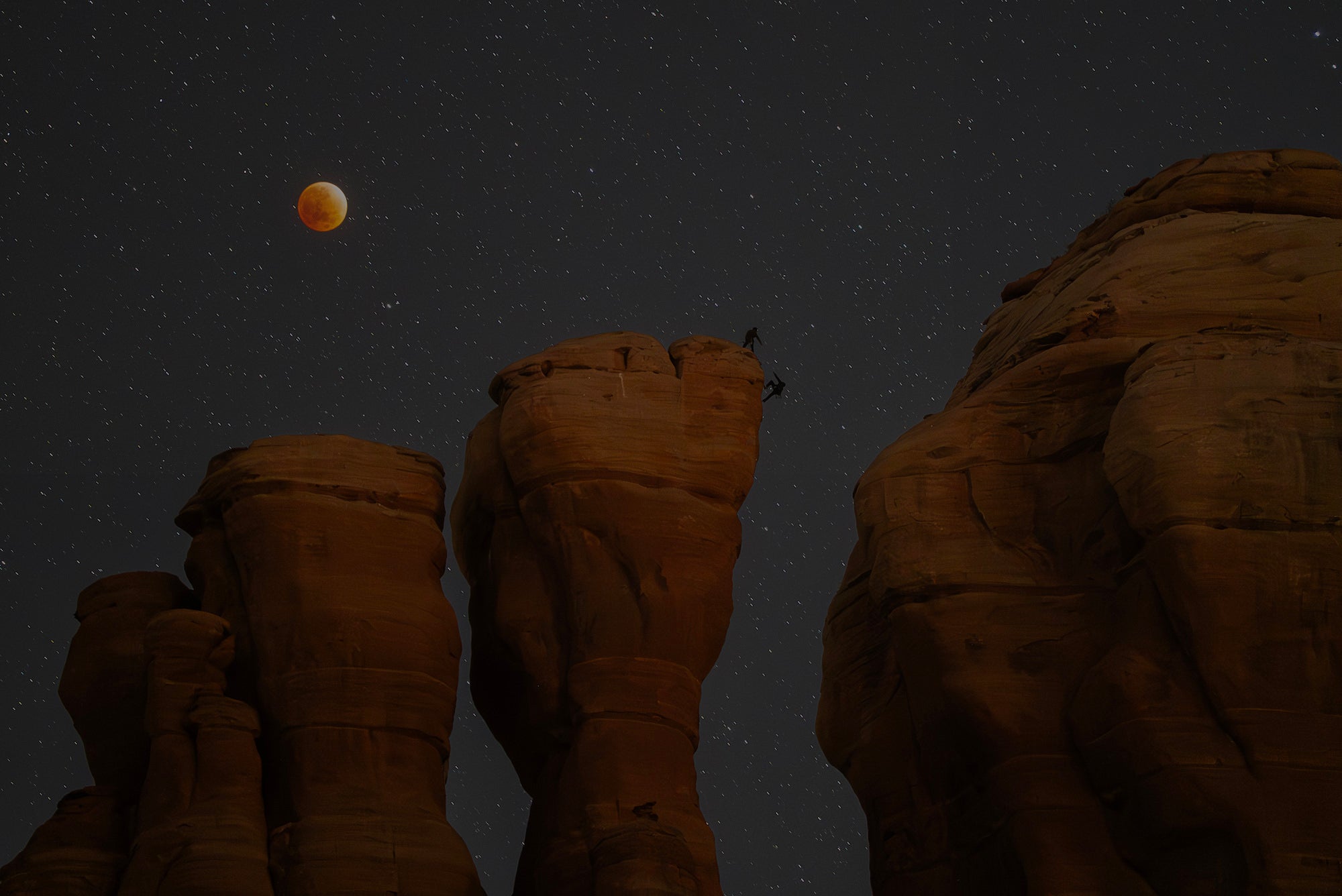 Blood moon over starry sky with tall sandstone towers and two climbers silhouetted on a summit. Blood moon over starry sky with tall sandstone towers and two climbers silhouetted on a summit.