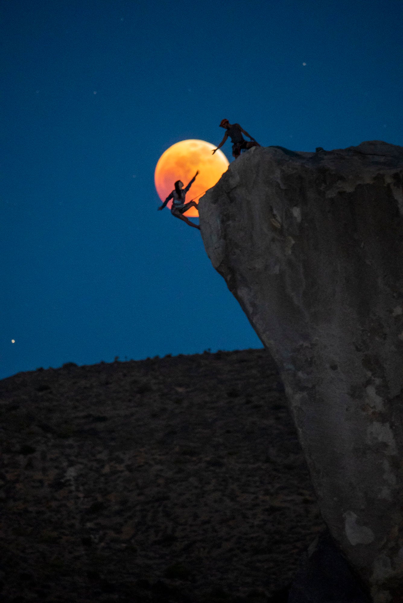 Two climbers silhouetted on a cliff edge as an orange-red eclipsed moon rises behind them. Two climbers silhouetted on a cliff edge as an orange-red eclipsed moon rises behind them.