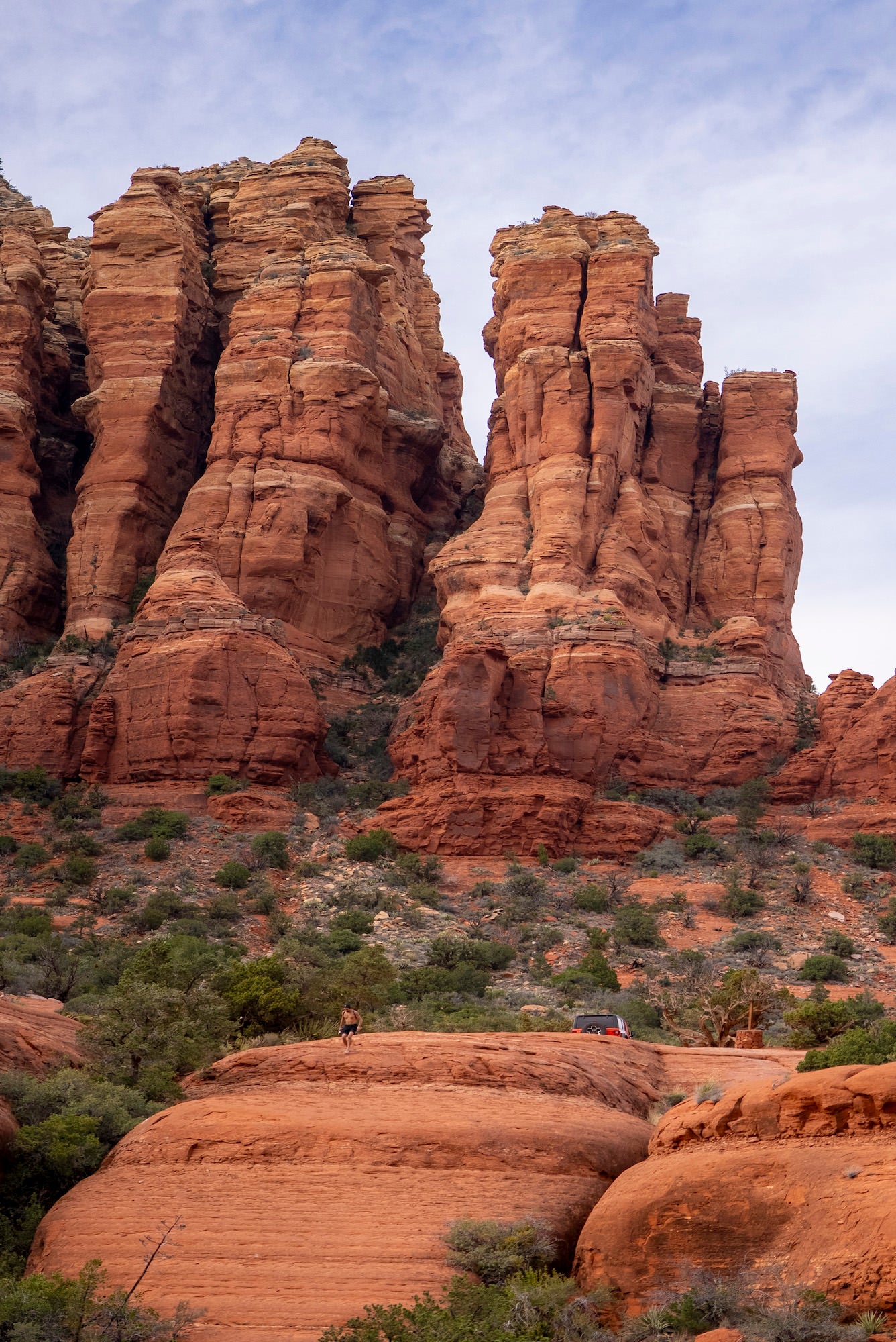 Wide view of Sedona red rock cliffs and desert vegetation with a hiker and parked vehicle in the foreground.