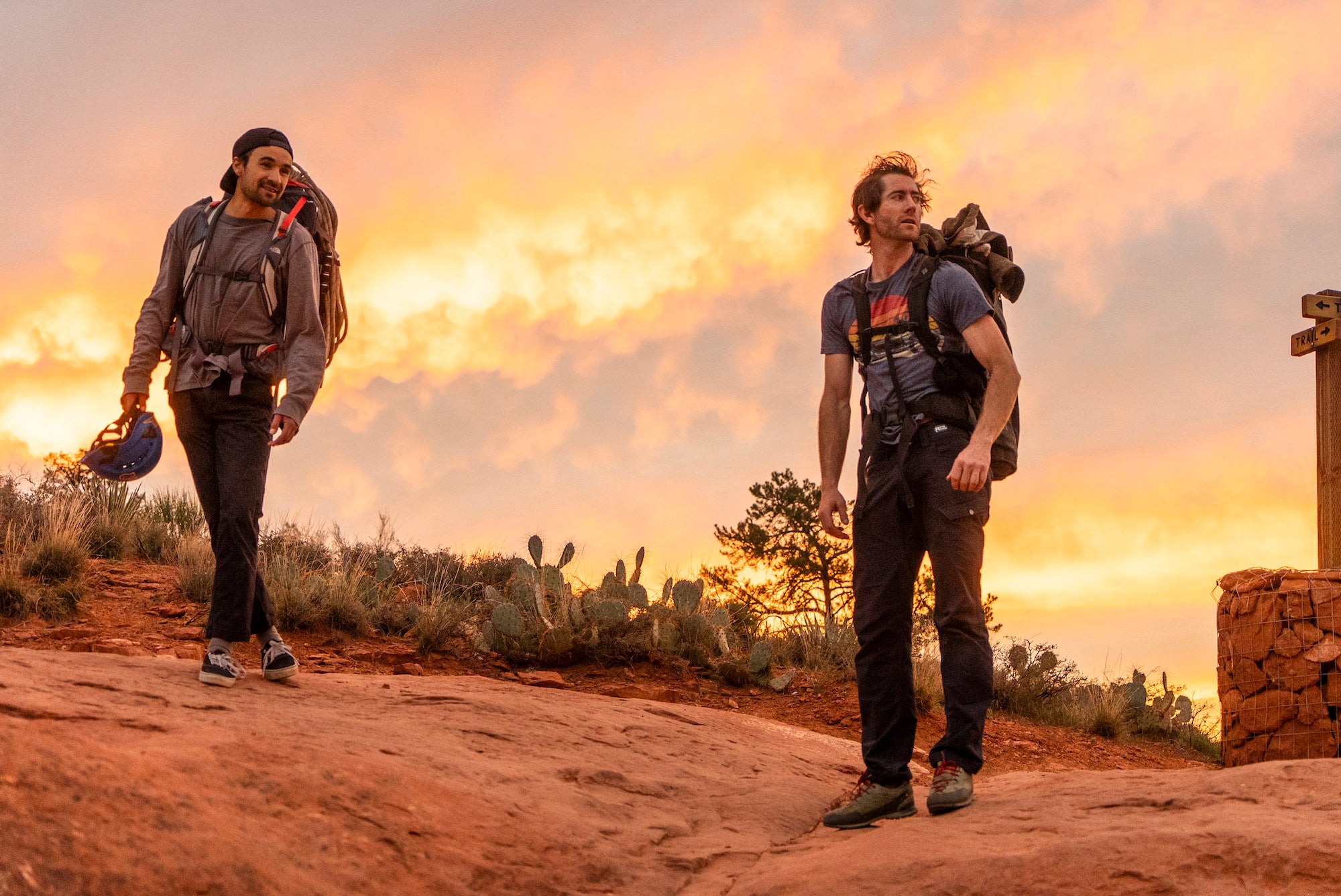 Two backpacked climbers walking at golden hour on a red rock trail beneath a glowing sky.
