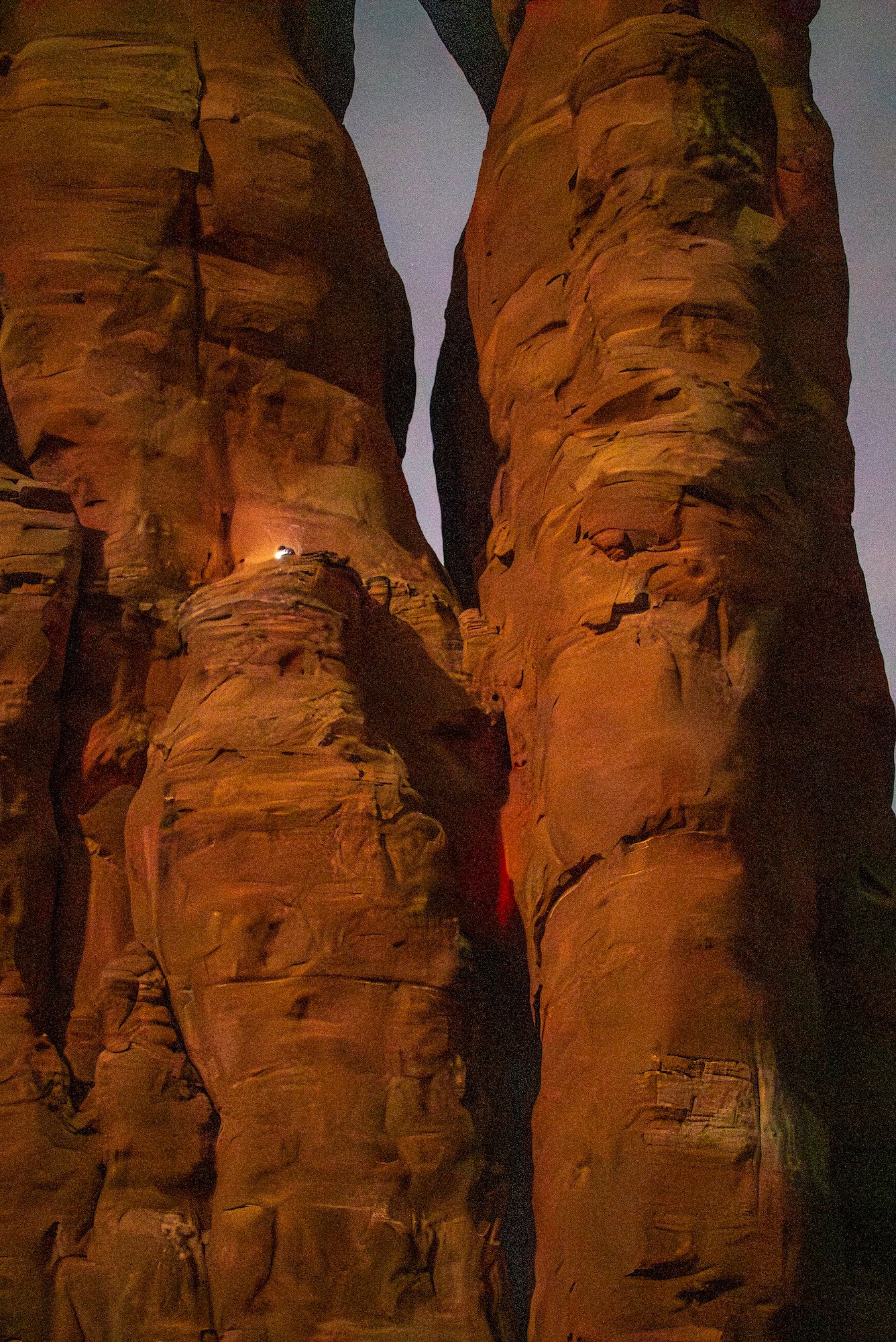 Headlamps illuminate a small ledge amid towering red rock walls during a nighttime climb.