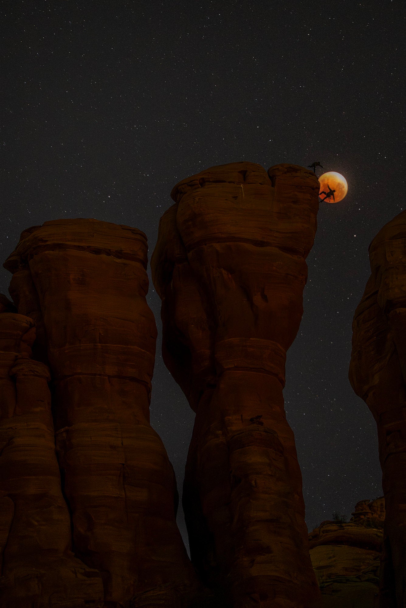 Lunar eclipse framed beside a sandstone tower as two climbers silhouette against the red moon and star field. Lunar eclipse framed beside a sandstone tower as two climbers silhouette against the red moon and star field.
