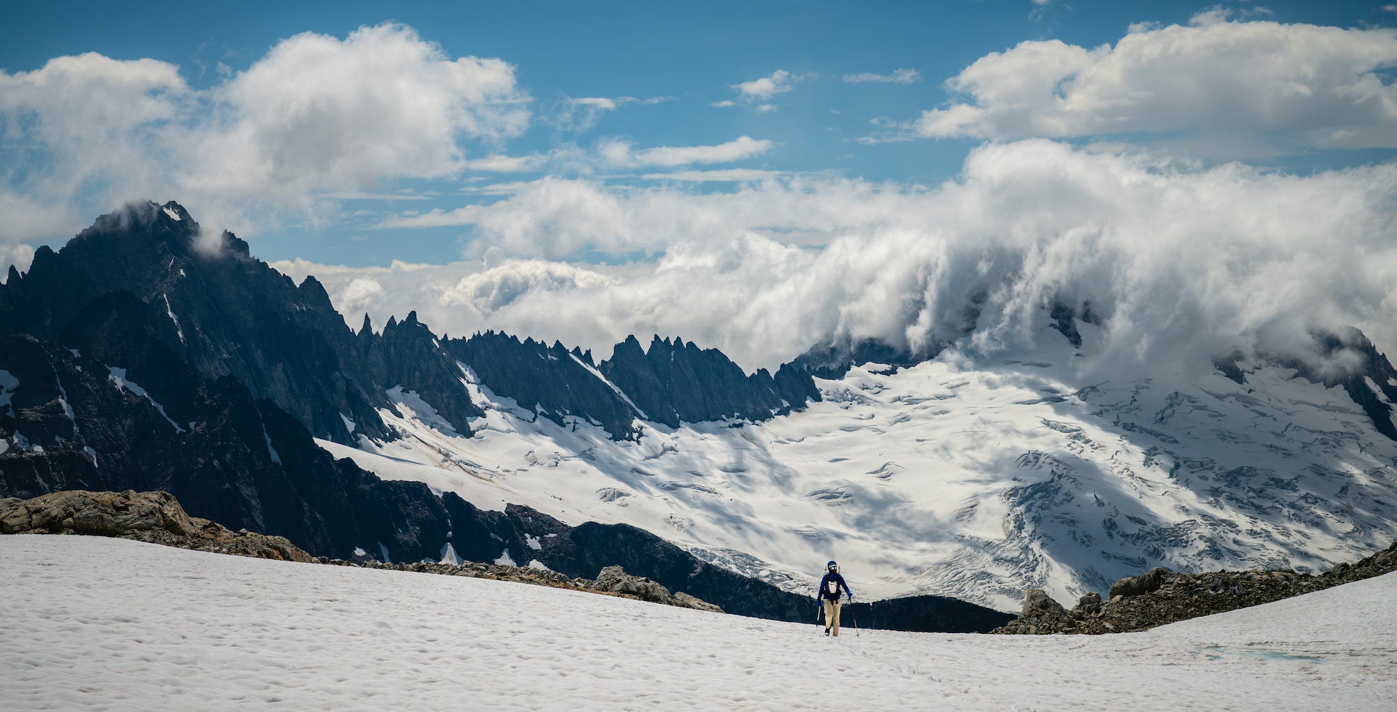 Climber crossing a broad snowfield beneath jagged North Cascades peaks as clouds spill over ridges under a bright sky.