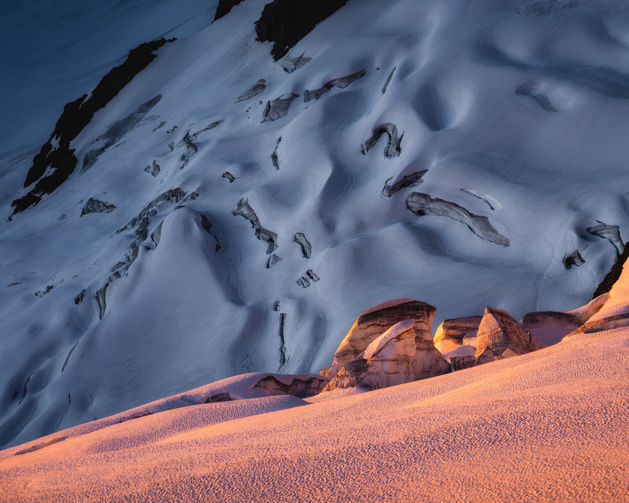 Close-up glacier scene with pink alpenglow on foreground seracs and blue shadowed crevasses beyond, highlighting warm&ndash;cool contrast.