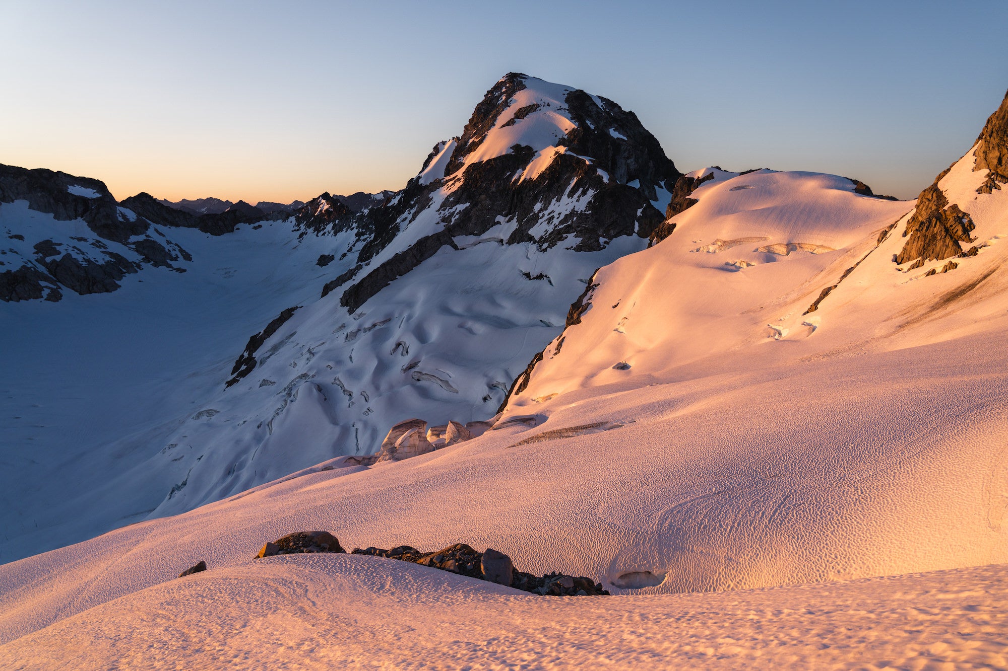 Sunrise washes a rolling glacier and adjacent peak in warm light while shadowed basins and distant ranges recede.