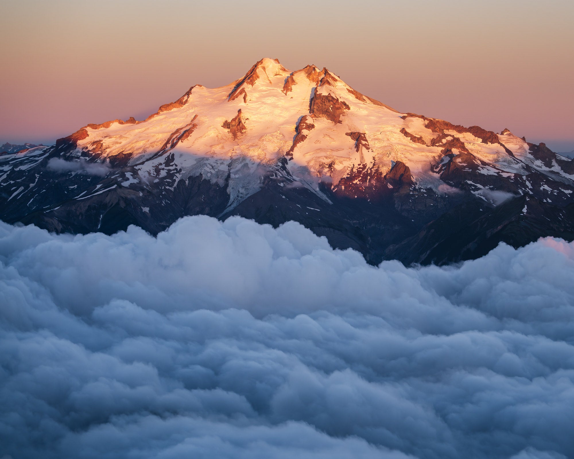 Glacier Peak (Dakobed) lit by sunrise above a sea of valley clouds, with soft pastel sky and snowfields glowing warm.