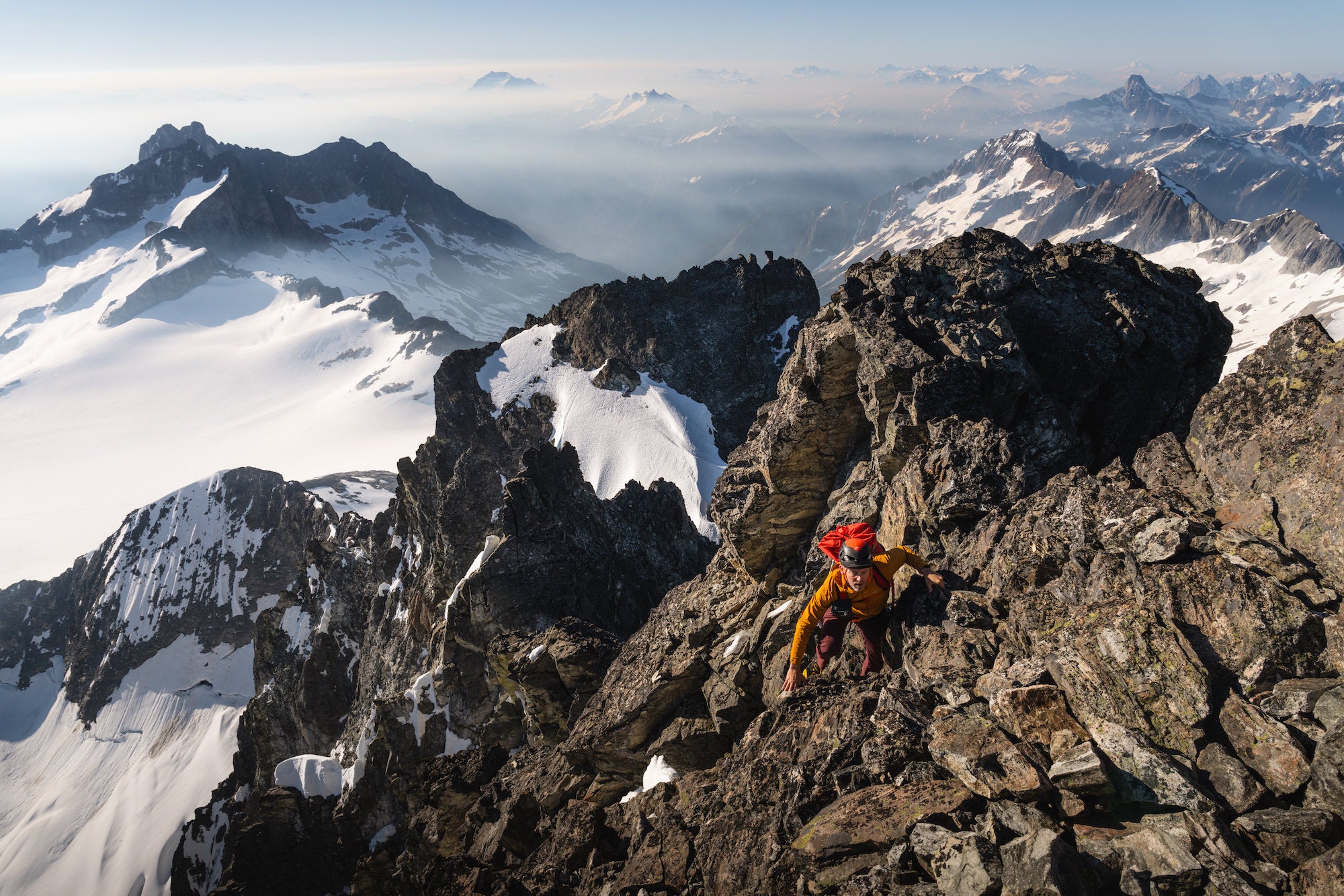 Climber scrambles steep blocky rock high above glaciers and distant volcanoes, with a sweeping North Cascades panorama.