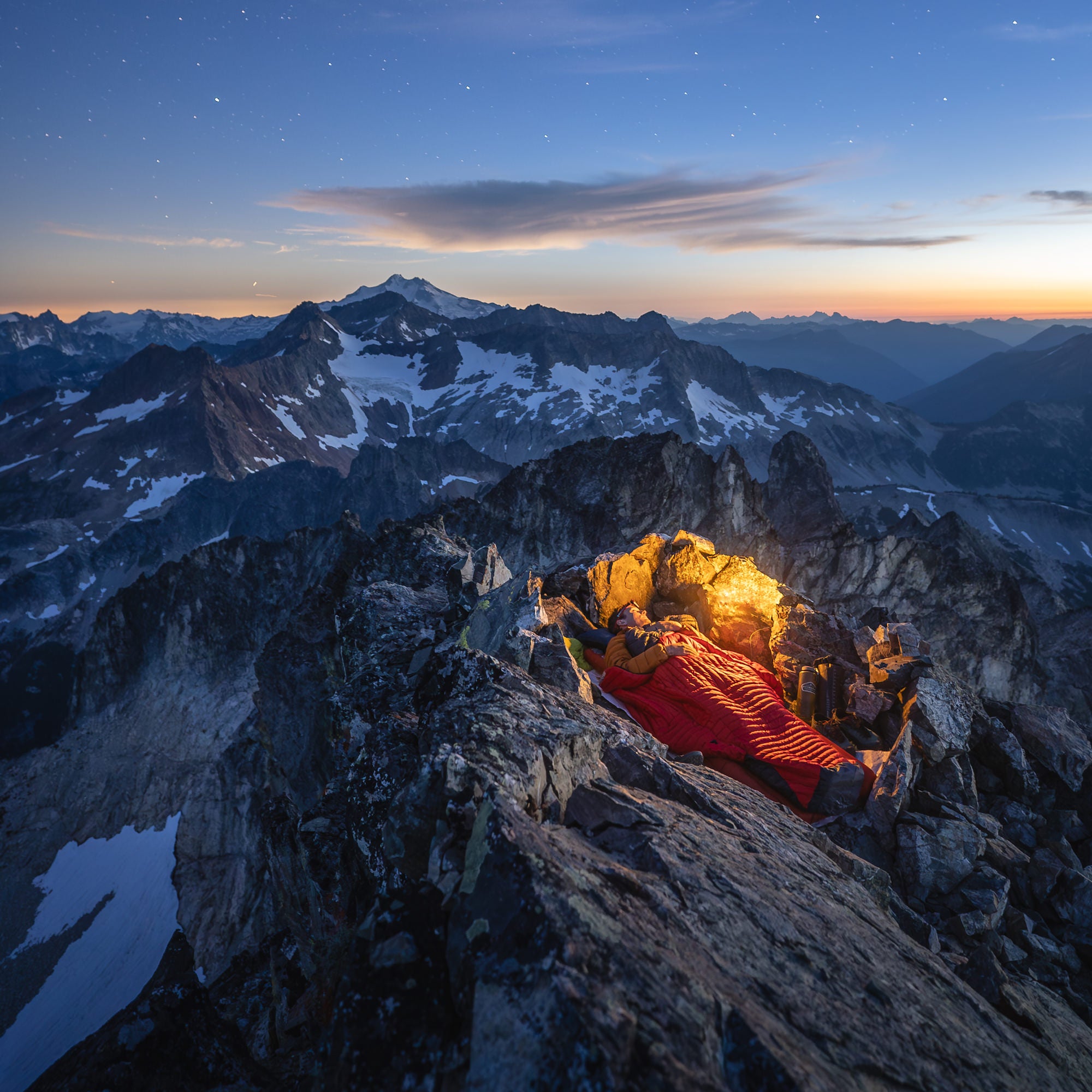 Solo summit bivy at twilight with a warm headlamp glow in a rock shelter under a starry sky and distant snowy peaks.