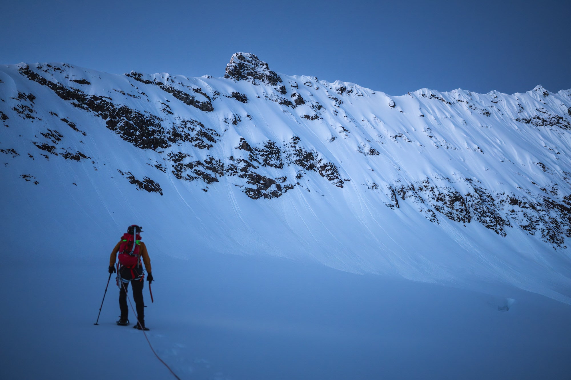 Pre-dawn blue hour on a glaciated slope as a roped climber approaches a steep, corniced ridge below dark rock.