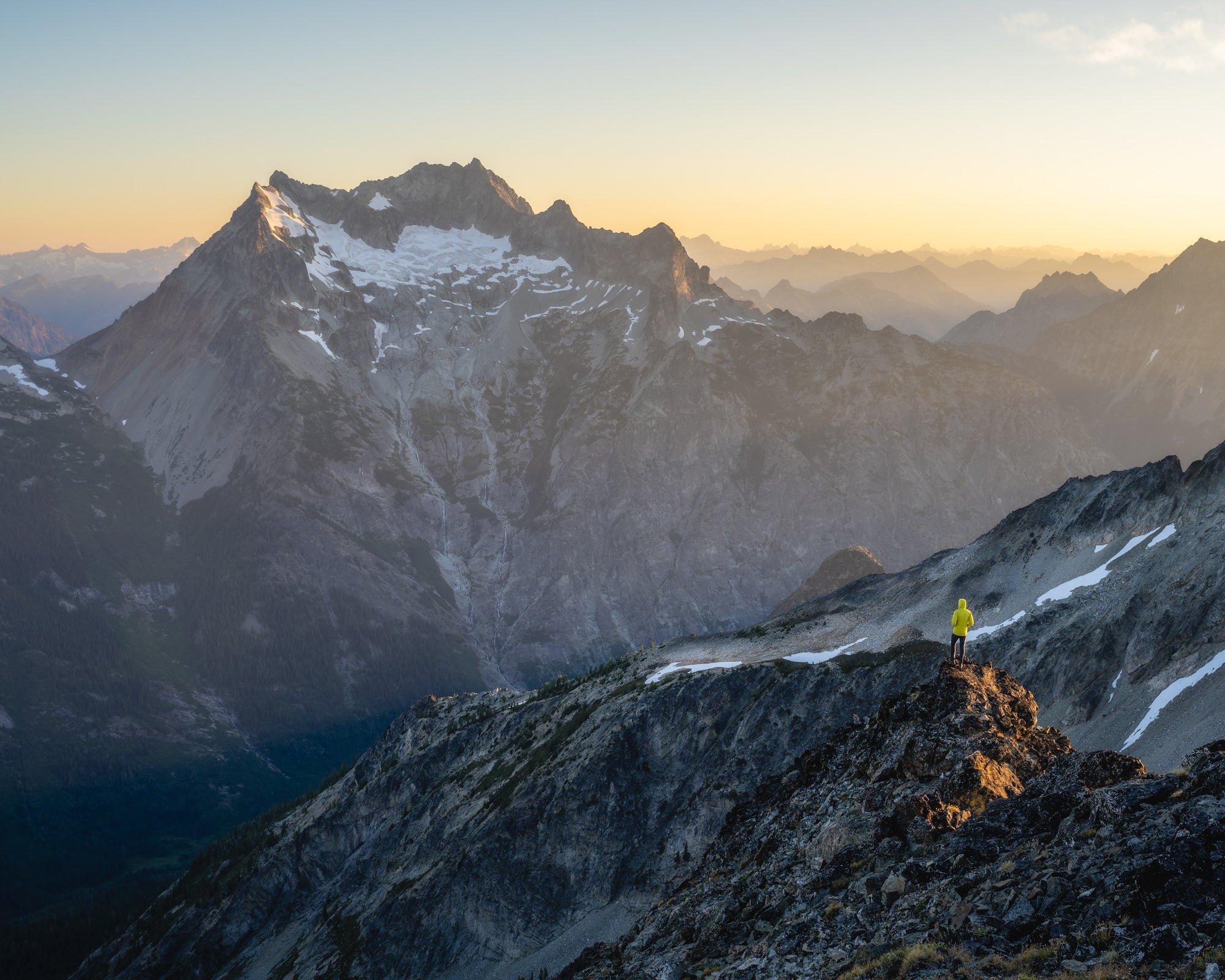 Figure in a yellow jacket stands on a sunlit ridge at golden hour, overlooking vast valleys and layered mountain ranges.