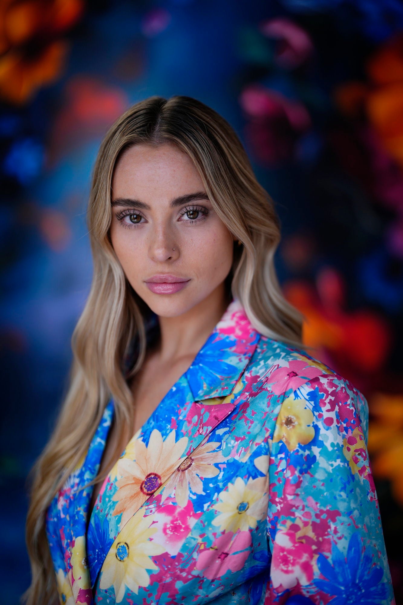 Portrait of a blonde woman in a bright floral blazer against a colorful blurred backdrop.
