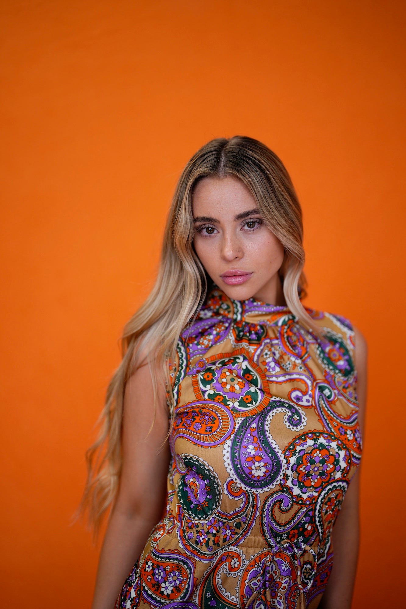 Blonde woman in a paisley-print sleeveless top posing against a bright orange backdrop.