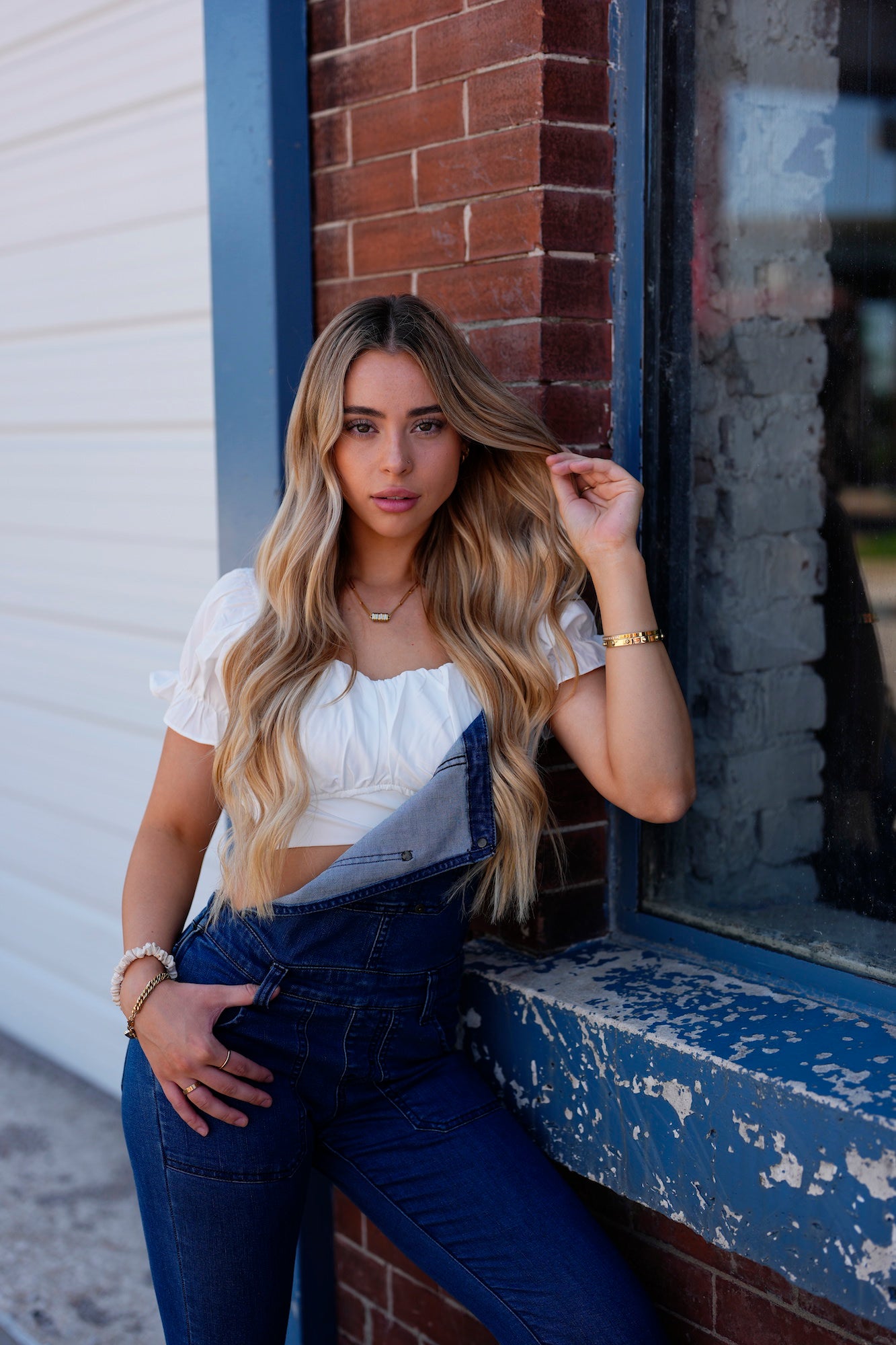Blonde woman in denim overalls and a white top leaning against a brick storefront window, touching her hair.